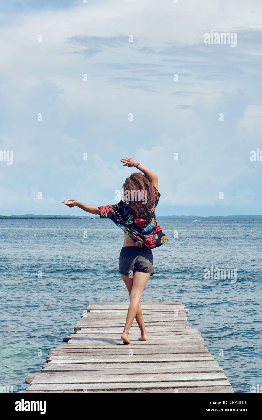 Vista posteriore di donne irriconoscibili in pantaloncini corti e top con lunghi capelli ondulati in piedi con le braccia a parte sulla fine del molo shabby godendo la brezza marina in sunli Foto Stock