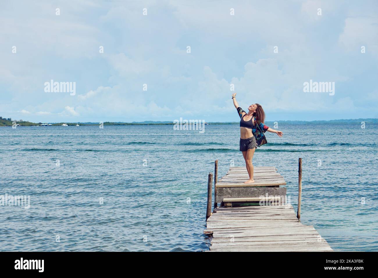 Donna lontana in pantaloncini corti e top con lunghi capelli ondulati in piedi con le braccia a parte sulla fine del molo shabby godendo la brezza marina alla luce del sole Foto Stock