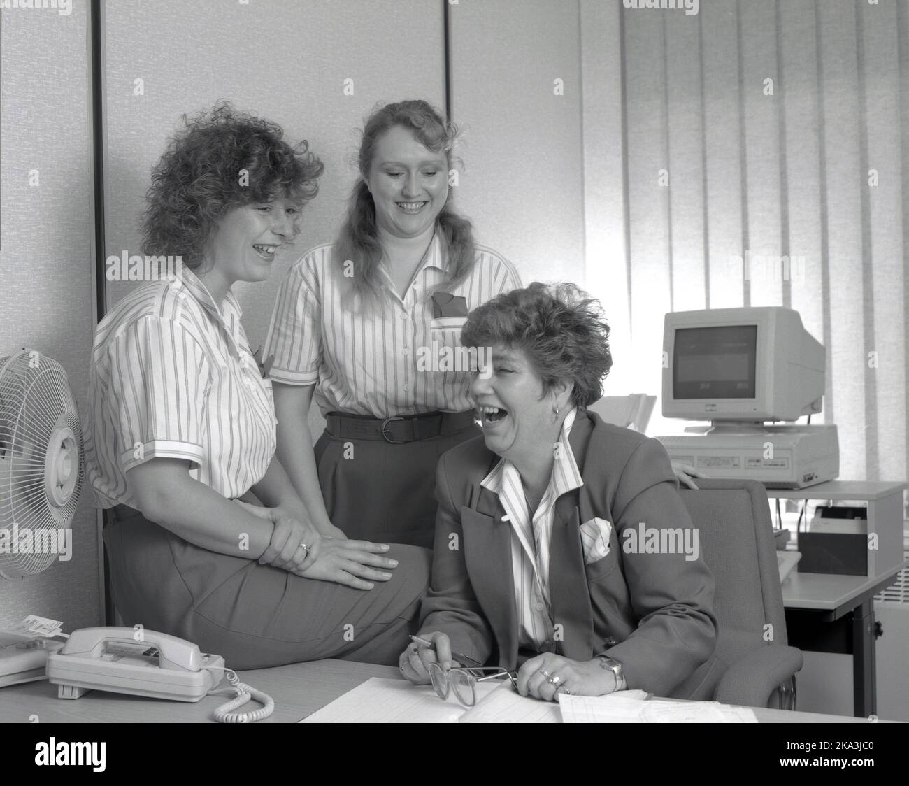 1990, storico, all'interno di un ufficio, tre donne del personale, tutte vestite con uniformi aziendali, che si divertono insieme, Inghilterra, Regno Unito. L'Office Manager indossa una giacca e si siede alla sua scrivania. Nella foto, situata su un'altra scrivania, un personal computer dell'epoca, costituito da un monitor VGA separato e un computer, un Amstrad PC2086, un modello prodotto dalla Gran Bretagna, un modello rilasciato nel 1988. Foto Stock