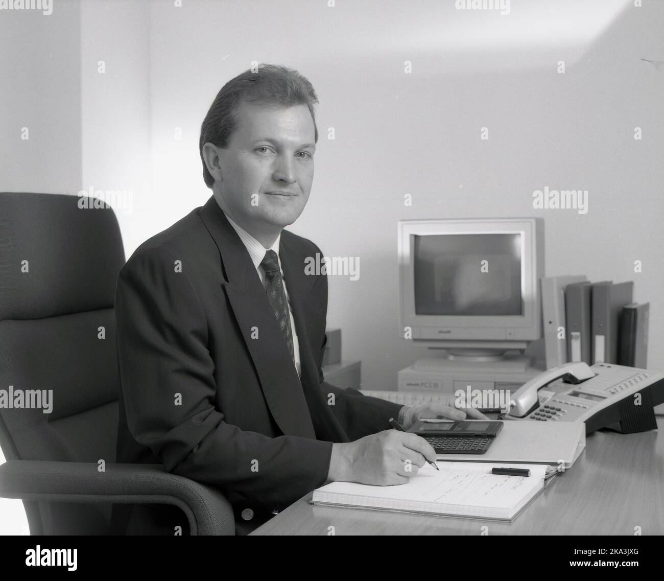1991, storico, un dirigente d'affari maschile adatto seduto alla sua scrivania con un notebook, cartella, telefono e organizzatore elettronico - noto anche come un assistente personale digitaal (PDA) - a portata di mano. Nella foto si vede anche un personal computer dell'epoca, un Olivetti PCS 286, con monitor VGA e tastiera. Lanciato nel 1990 da Olivetti, uno dei principali produttori italiani di computer, questo PC desktop a ingombro ridotto è stato progettato per i mercati domestici e aziendali, con una memoria R.A.M di 1MB GB e un'unità floppy disk da 3,5'. Foto Stock