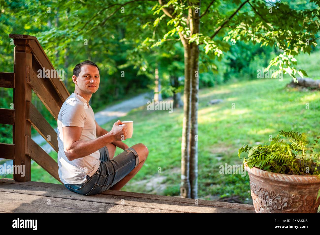 Portico di casa d'estate con l'uomo seduto su gradini di casa di fronte o sul retro cortile mattina cabina di legno cottage bere caffè o tè da tazza guardando indietro Foto Stock