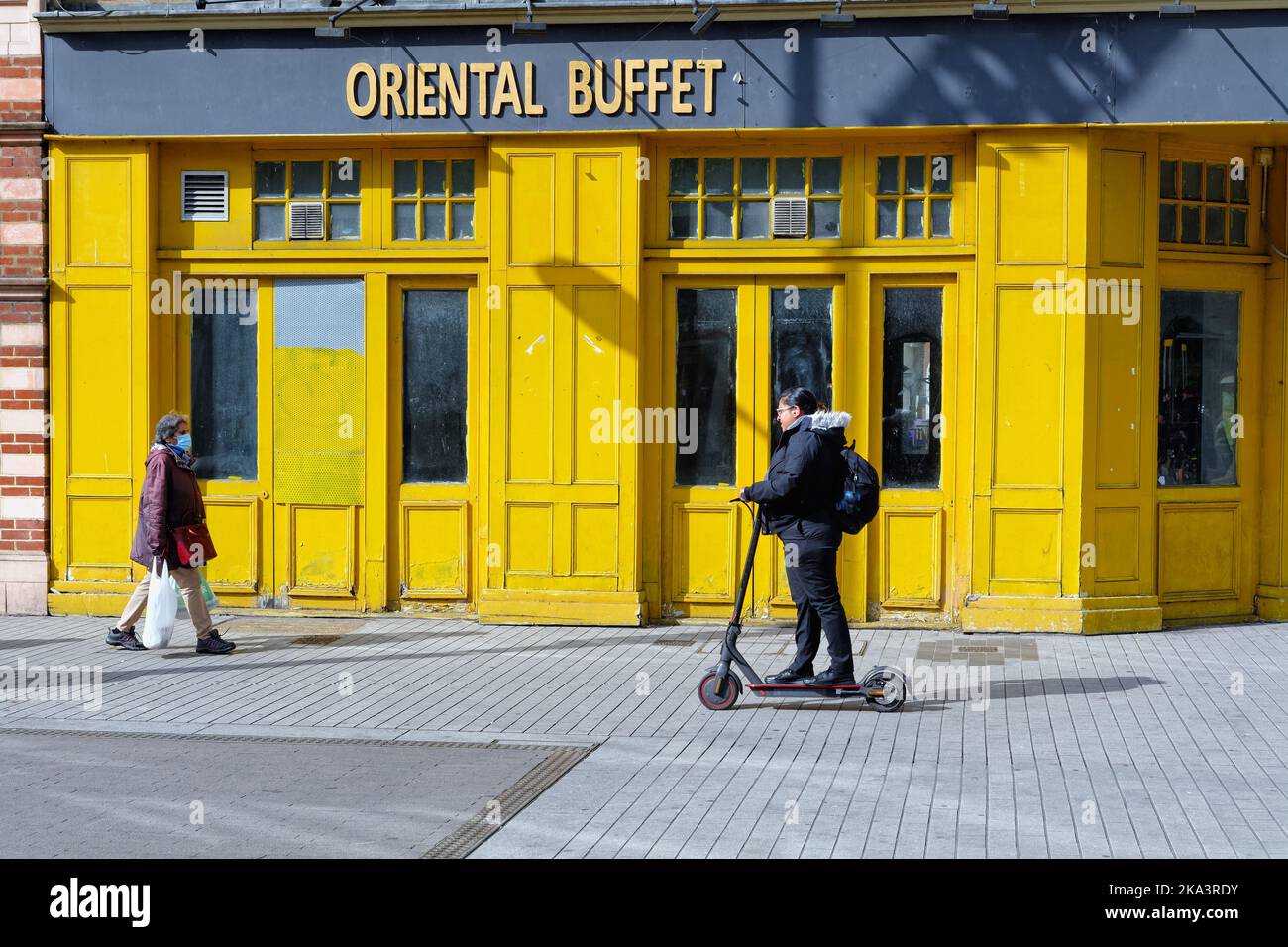 Una giovane ragazza asiatica in sella a uno scooter elettrico sul marciapiede all'esterno di un colorato e chiuso 'Oriental buffet' a Hounslow High Street West London Inghilterra UK Foto Stock