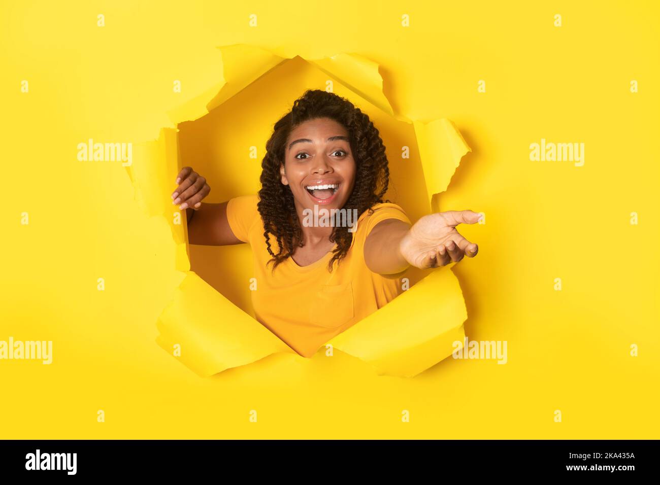 Excited African Lady che si allunga mano attraverso il foro su sfondo giallo Foto Stock