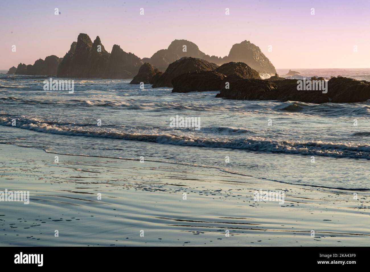 All'alba o al tramonto sulla costa dell'Oregon, con onde che si lavano su rocciatori ruvidi per raggiungere la spiaggia sabbiosa. Foto Stock