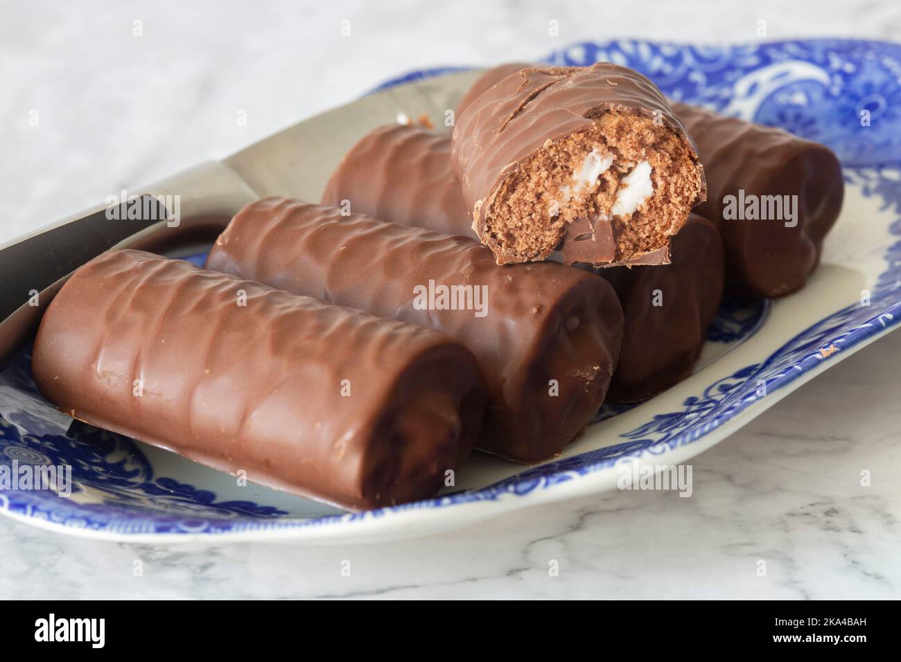 Rotoli di spugna al cioccolato individuali su un piatto blu e bianco a motivi geometrici con un coltello da portata. Su uno sfondo di marmo bianco Foto Stock