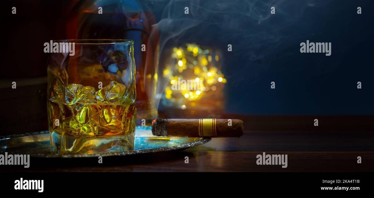 un bicchiere di whisky con ghiaccio su un tavolo di legno, un sigaro cubano fumante e una bottiglia di whisky in un'atmosfera fumosa di un bar notturno Foto Stock