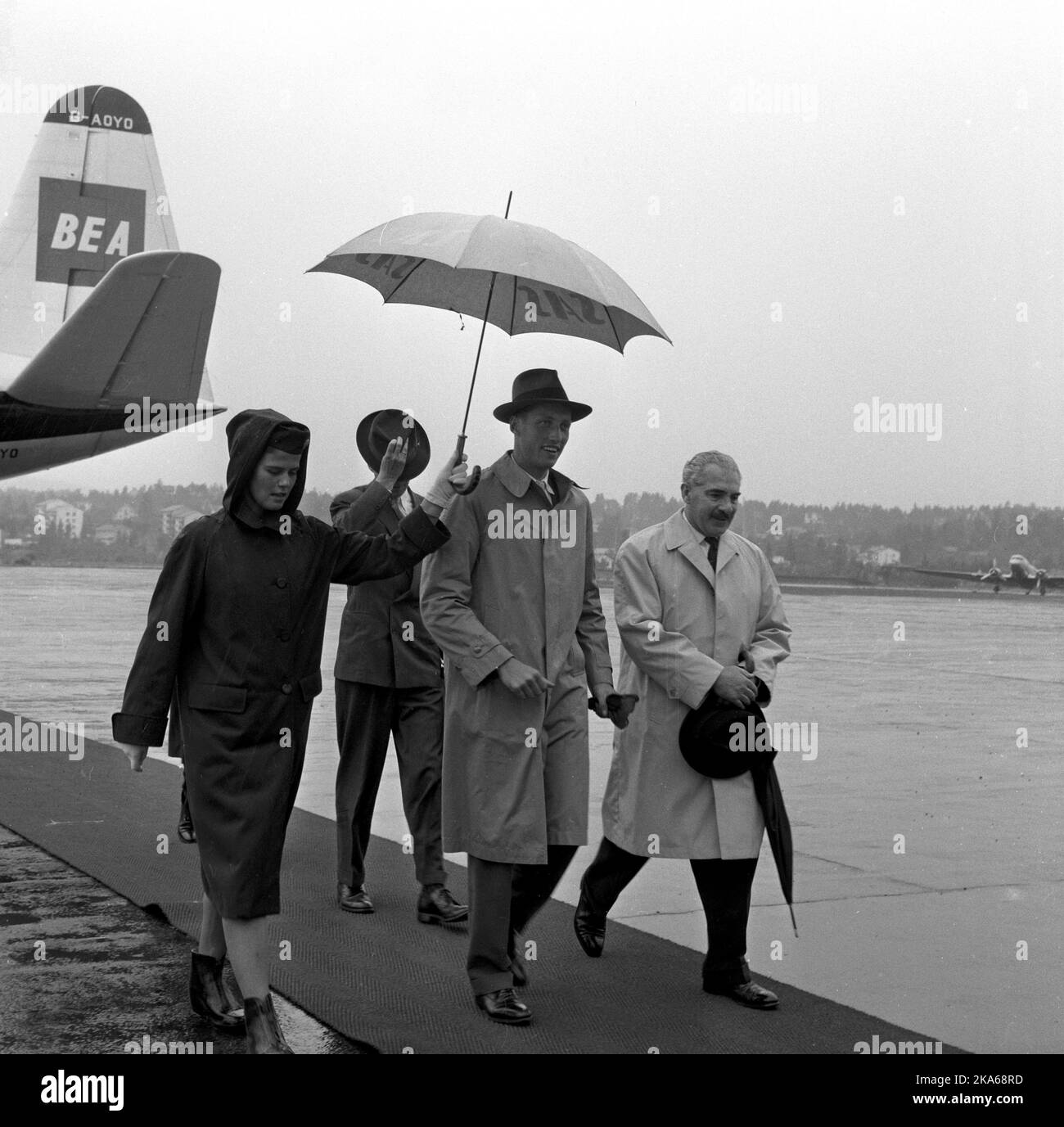 Aftenposten. Fornebu , Oslo 19610709 : VT ACCOGLIE Øsende le piogge quando le LP rosse furono lanciate per il principe ereditario Harald , che venne con l'aereo BEA da Londra. Il suo college vårsemester PA Balliol è finito. In caso contrario, pa fotografa la hostess SAS Aina Hansen (a sinistra) con ombrello sopra il principe della corona , l'adiuvante del principe della corona , il comandante gennaio Ørner (a retro) e il presidente LHG Kent , capo della British European Airways in Norvegia . Foto: Brynjulv Aartun / Aftenposten *** FOTO IKKE BILDEBEHANDLET *** Foto Stock