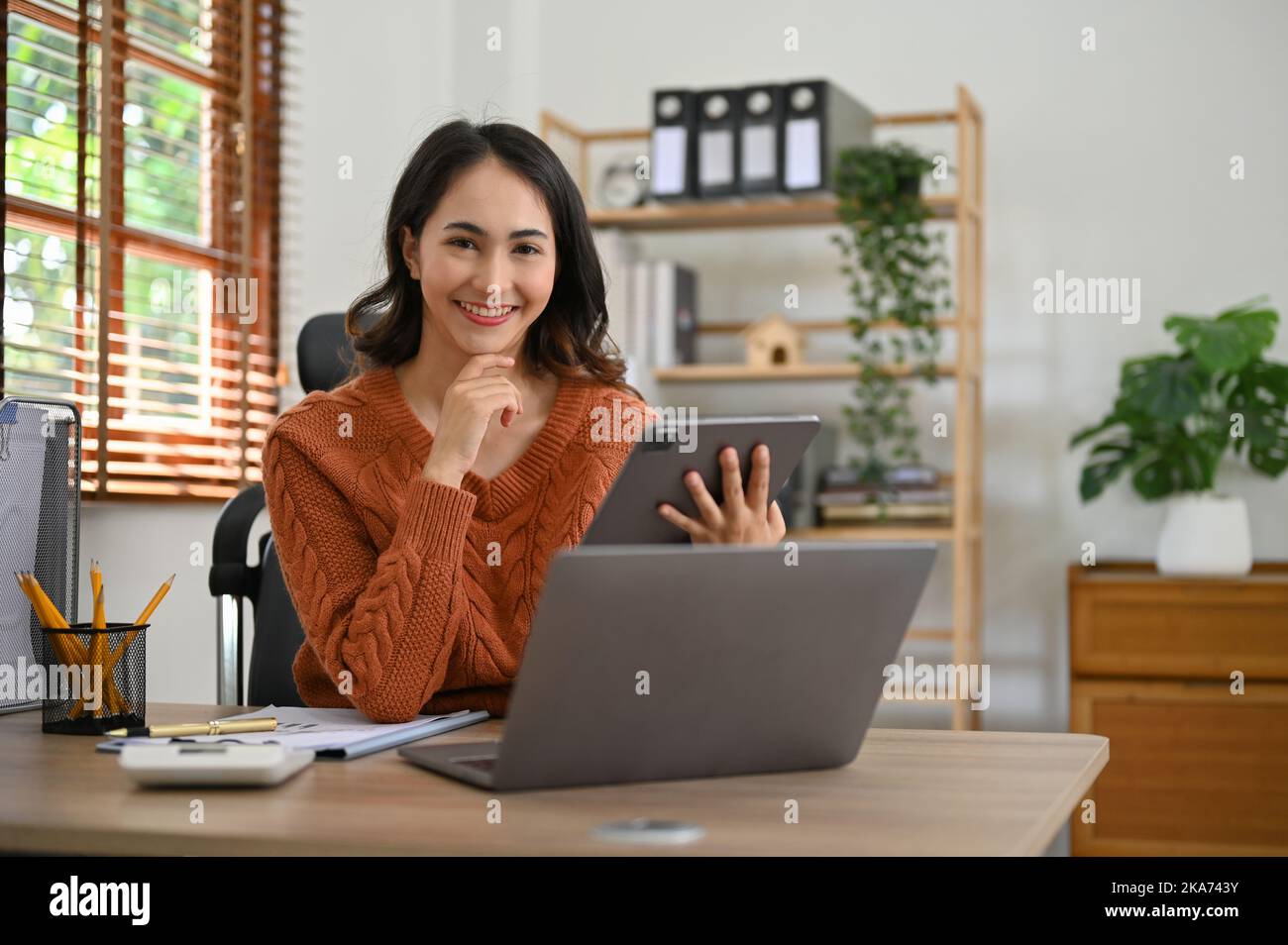 Affascinante donna d'affari asiatica millenaria in abiti casual seduto alla sua scrivania ufficio, tenendo un tablet portatile, mano sul mento, sorridendo e guardando ca Foto Stock