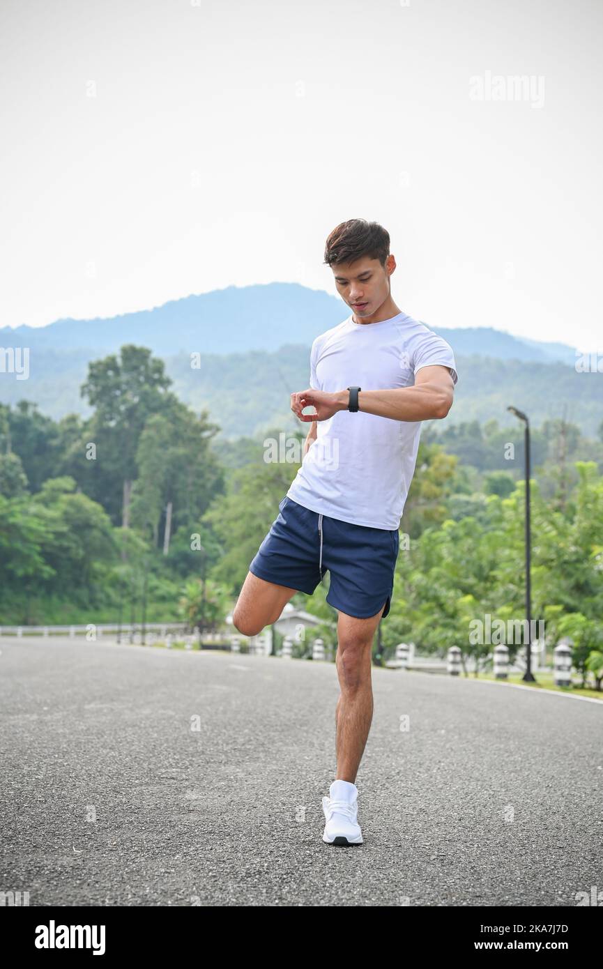 Ritratto, bello e sano giovane asiatico in abbigliamento sportivo allungando le gambe, guardando il suo orologio sportivo intelligente, contando le sue calorie e a. Foto Stock