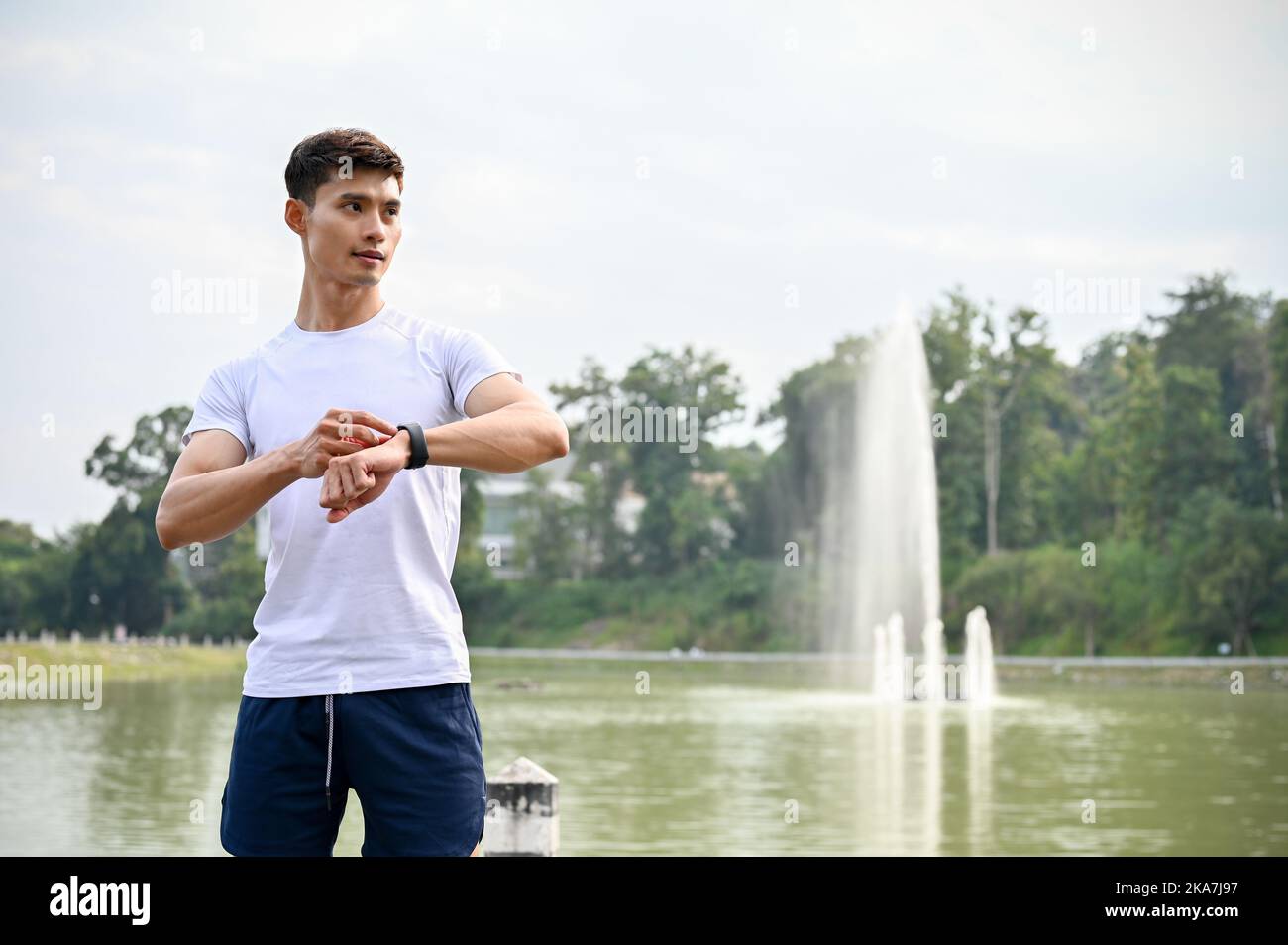 Bel giovane atletico asiatico in abbigliamento sportivo con il suo smartwatch, in piedi all'esterno, contando le calorie, preparandosi a correre. Attività sana con t Foto Stock
