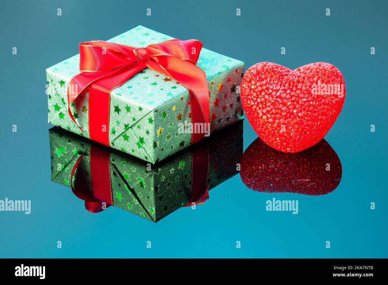 Confezione regalo verde singola con cuore rosso su superficie riflettente blu. Confezione regalo avvolta in carta lucida verde con nastro rosso Foto Stock