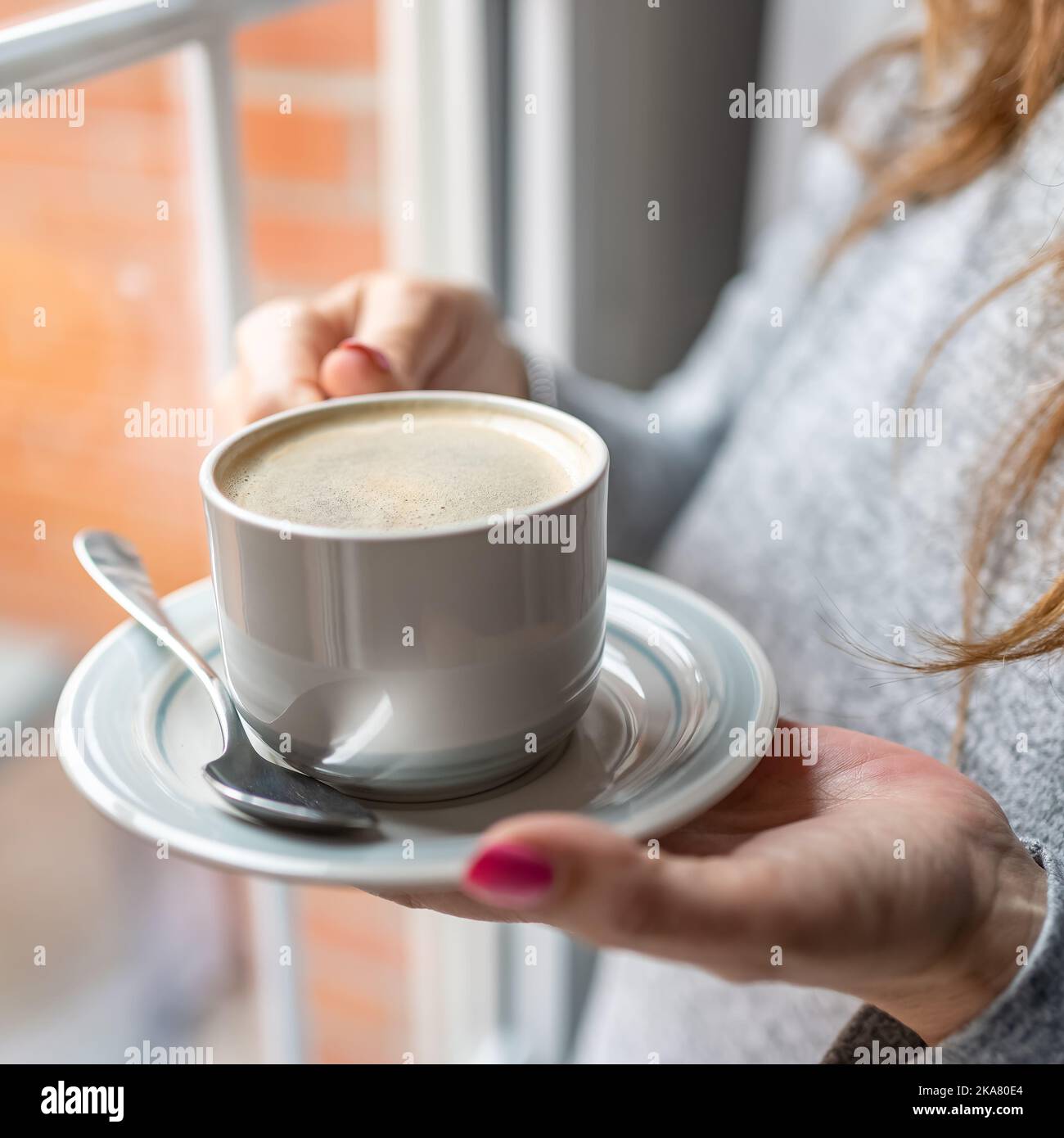 Le mani delle donne che tengono una tazza di caffè vicino alla finestra della loro casa. Foto Stock