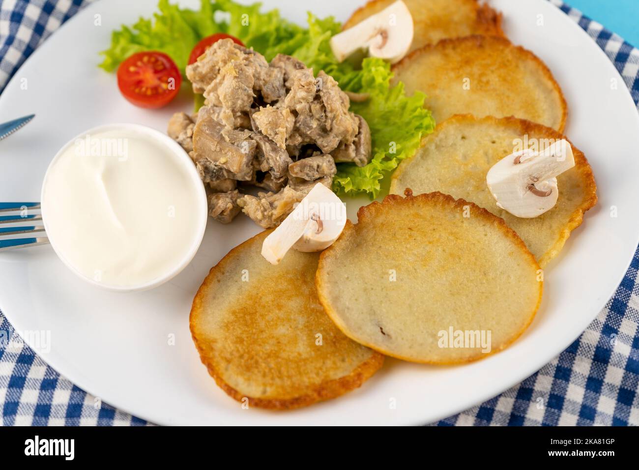 Deruny - frittelle di patate con funghi, champignons, panna acida su un piatto bianco. Pomodori ciliegini, insalata. Ristorante che serve su uno sfondo blu, tovagliolo a scacchi, forchetta, coltello. Vista dall'alto, spazio di copia Foto Stock