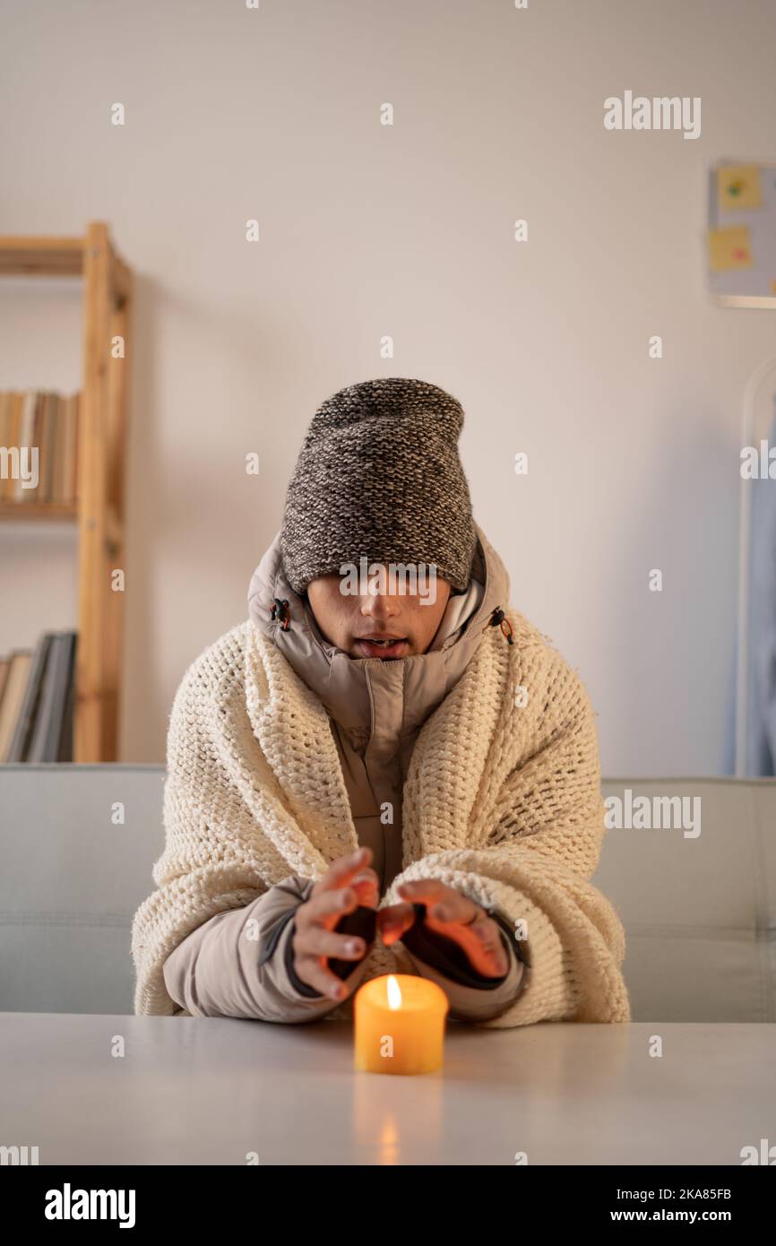 Un giovane gelatore vestito con un caldo cappello invernale e una giacca si siede a casa a un tavolo e riscalda le mani da una candela che brucia. Concetto di assenza di riscaldamento Foto Stock