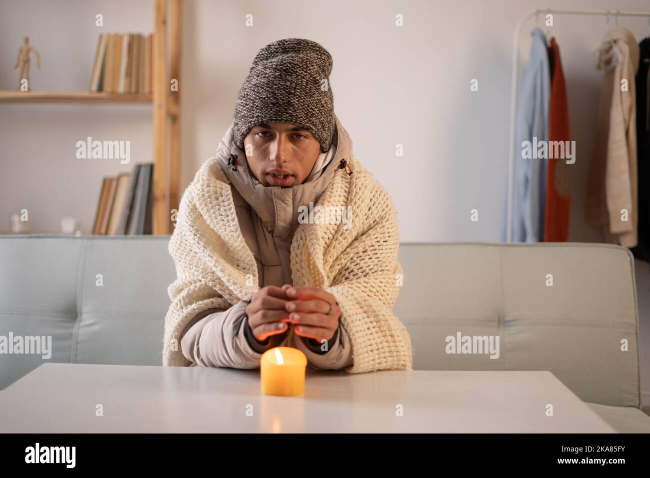 Un giovane gelatore vestito con un caldo cappello invernale e una giacca si siede a casa a un tavolo e riscalda le mani da una candela che brucia. Concetto di assenza di riscaldamento Foto Stock