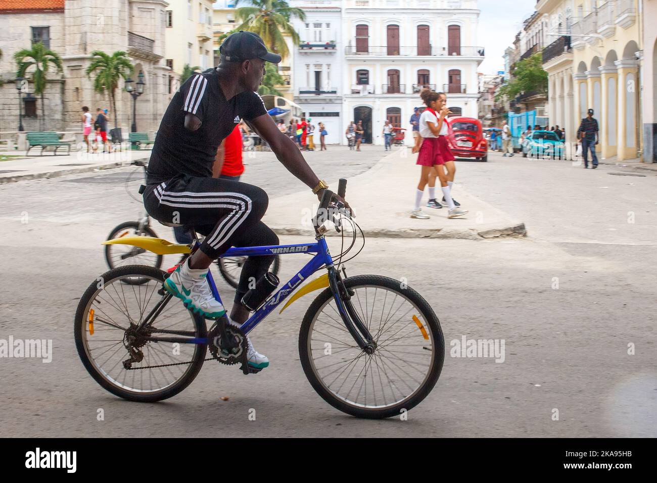 Un uomo cubano afro-caraibico che vive con una disabilità cavalca una bicicletta mentre i pionieri degli studenti in uniforme camminano sullo sfondo Foto Stock