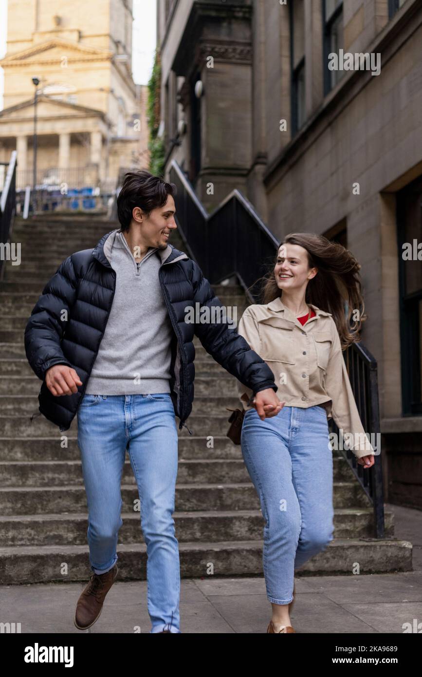 Una giovane coppia che trascorre la giornata a Newcastle upon Tyne insieme. Stanno tenendo le mani e correndo attraverso la città con i passi dietro di loro. Foto Stock