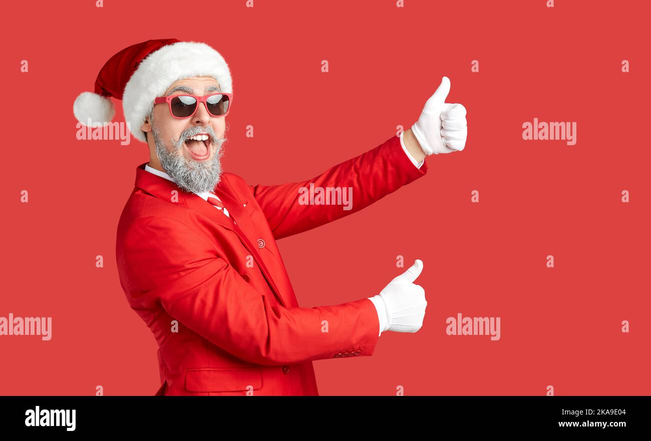 Uomo felice in cappello di Natale e occhiali da sole in piedi su sfondo rosso e dando pollici in su Foto Stock