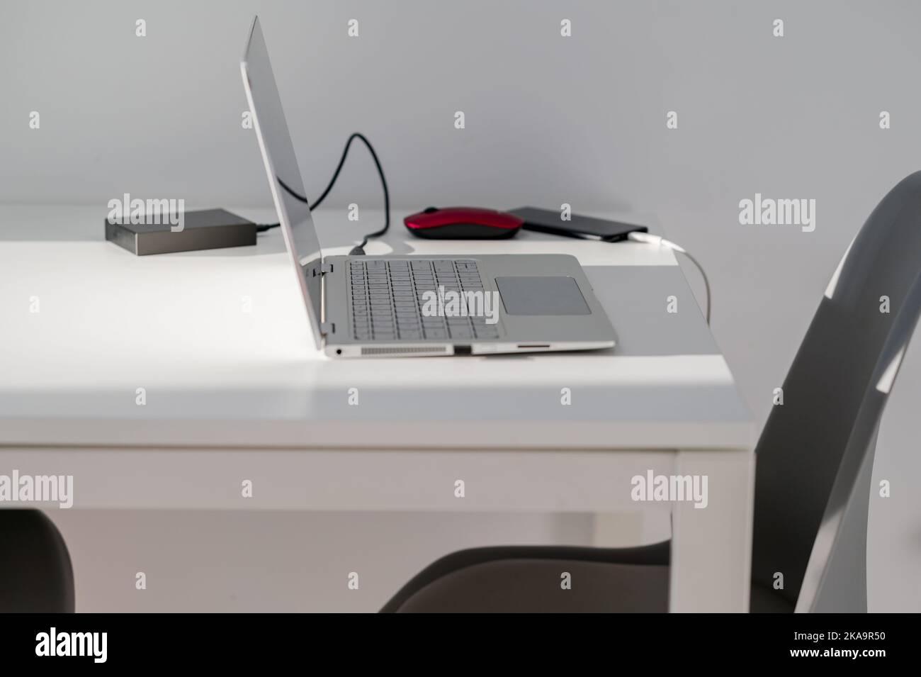 Un allestimento a casa con tavolo bianco con computer portatile, mouse rosso, disco rigido e telefono con sedie grigie e parete bianca. Lavoro a distanza. Foto Stock