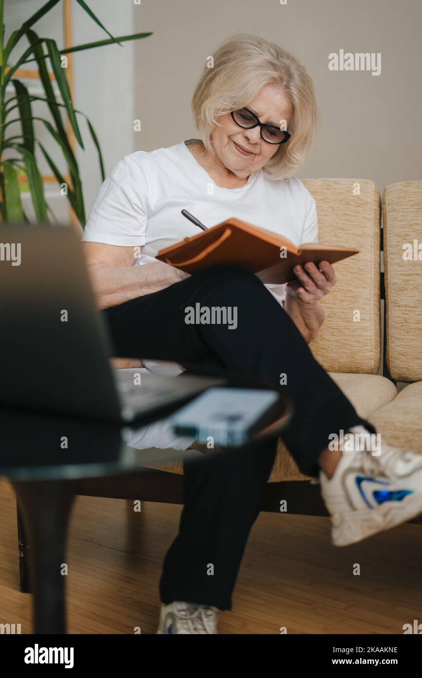 Donna anziana felice che guarda il webinar di formazione online utilizzando il notebook, il lavoro a distanza, la scrittura di note. Donna anziana che usa il notebook mentre si siede sul divano Foto Stock