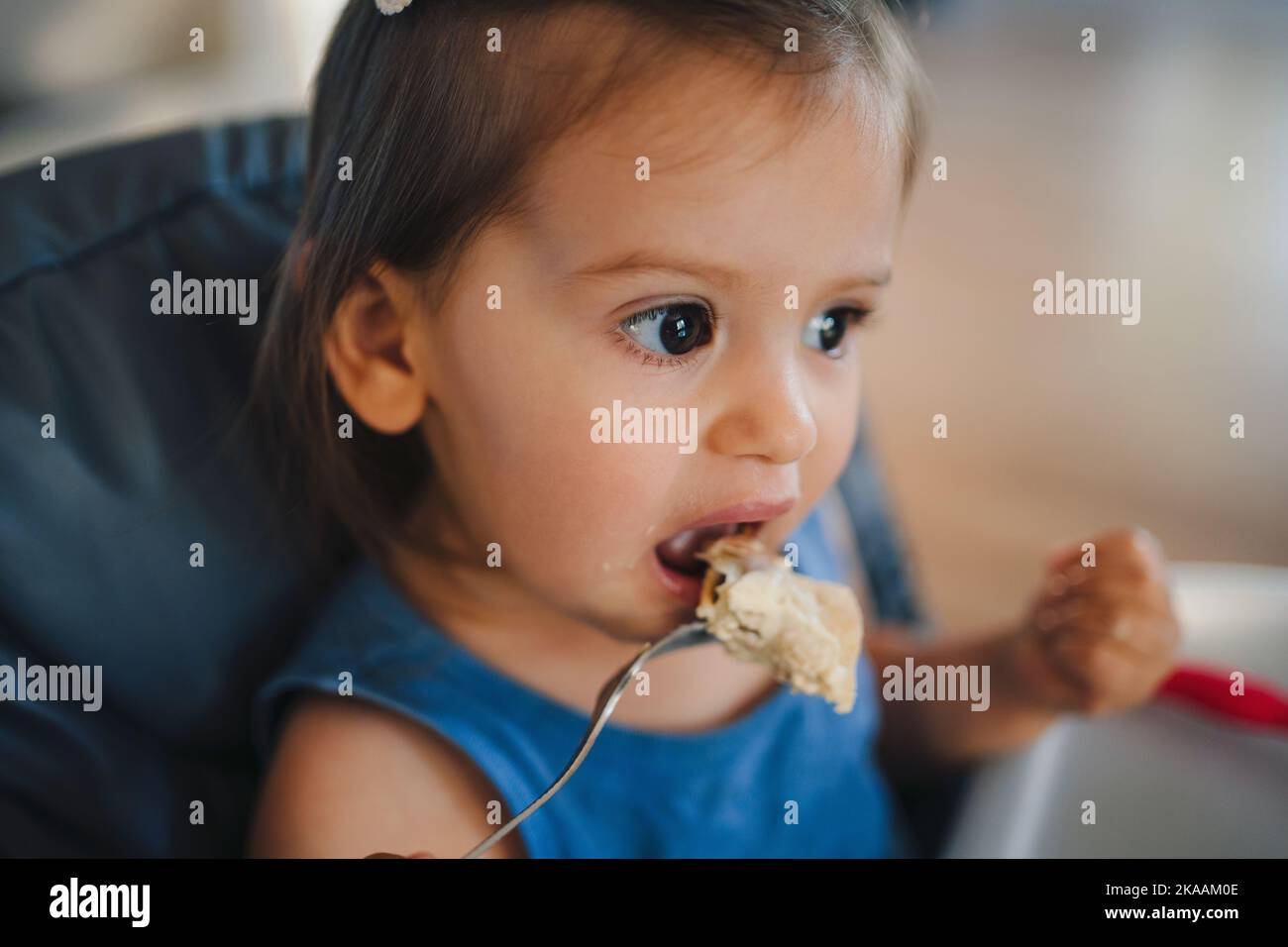 Adorabile ragazza caucasica seduta in seggiolone mangiare la sua cena con forchetta. Il bimbo mangia in modo indipendente. Un vero e proprio momento autentico. Primo Foto Stock