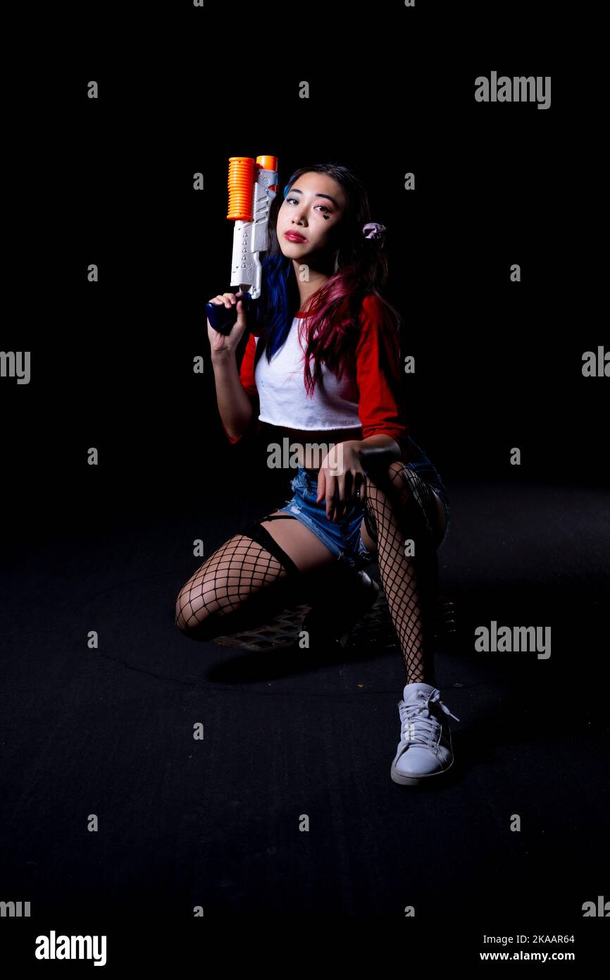 Giovane donna che tiene la pistola d'acqua vestita in costume di Harlequin su una strada di Asphalt Foto Stock