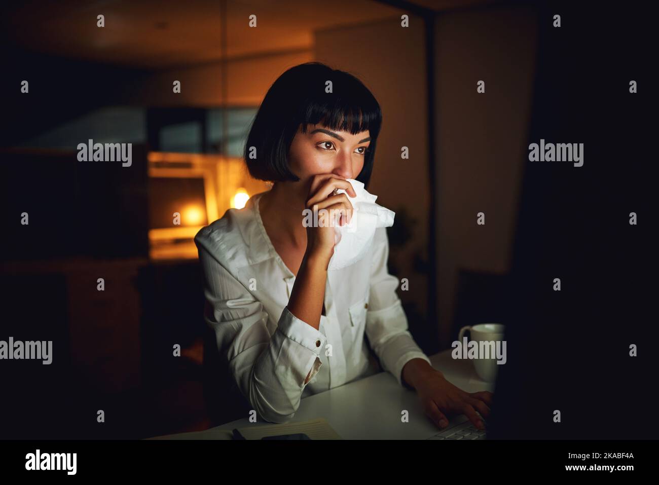 Le sere più tardi brindano con il suo sistema immunitario, una giovane donna d'affari che soffia il naso durante la notte tarda al lavoro. Foto Stock