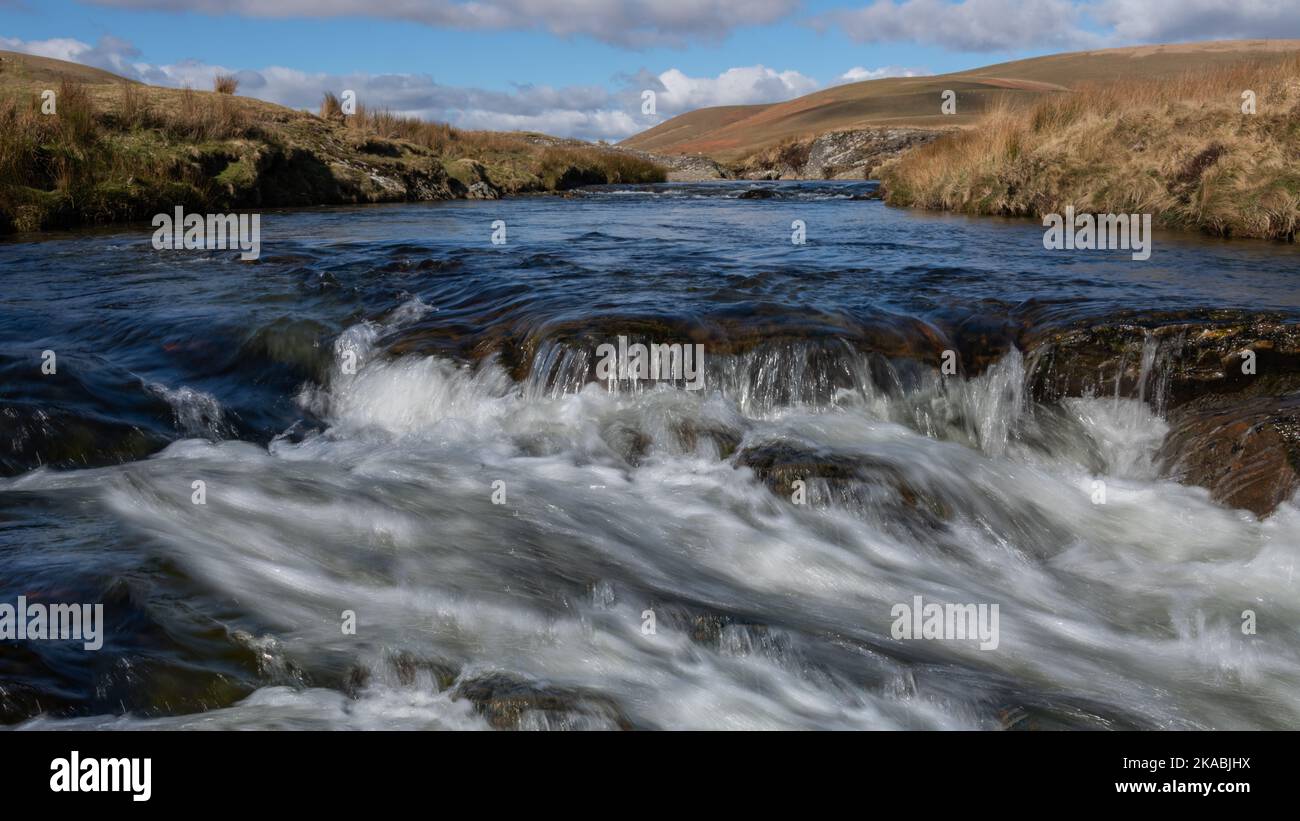 Un'immagine di basso livello che guarda su un piccolo fiume verso colline lontane. Mostra piccole rapide di acqua bianca in primo piano Foto Stock
