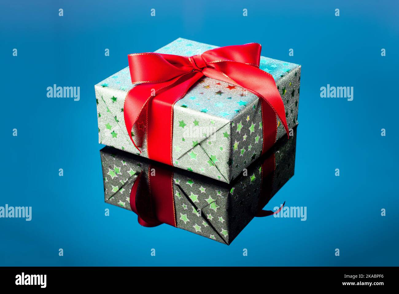 Confezione regalo verde singola con arco a nastro rosso su superficie riflettente blu. Confezione regalo confezionata in carta lucida verde con nastro rosso da vicino Foto Stock