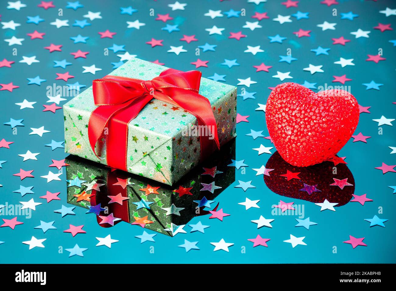 Confezione regalo verde singola con cuore rosso su superficie riflettente blu. Confezione regalo avvolta in carta lucida verde con nastro rosso Foto Stock