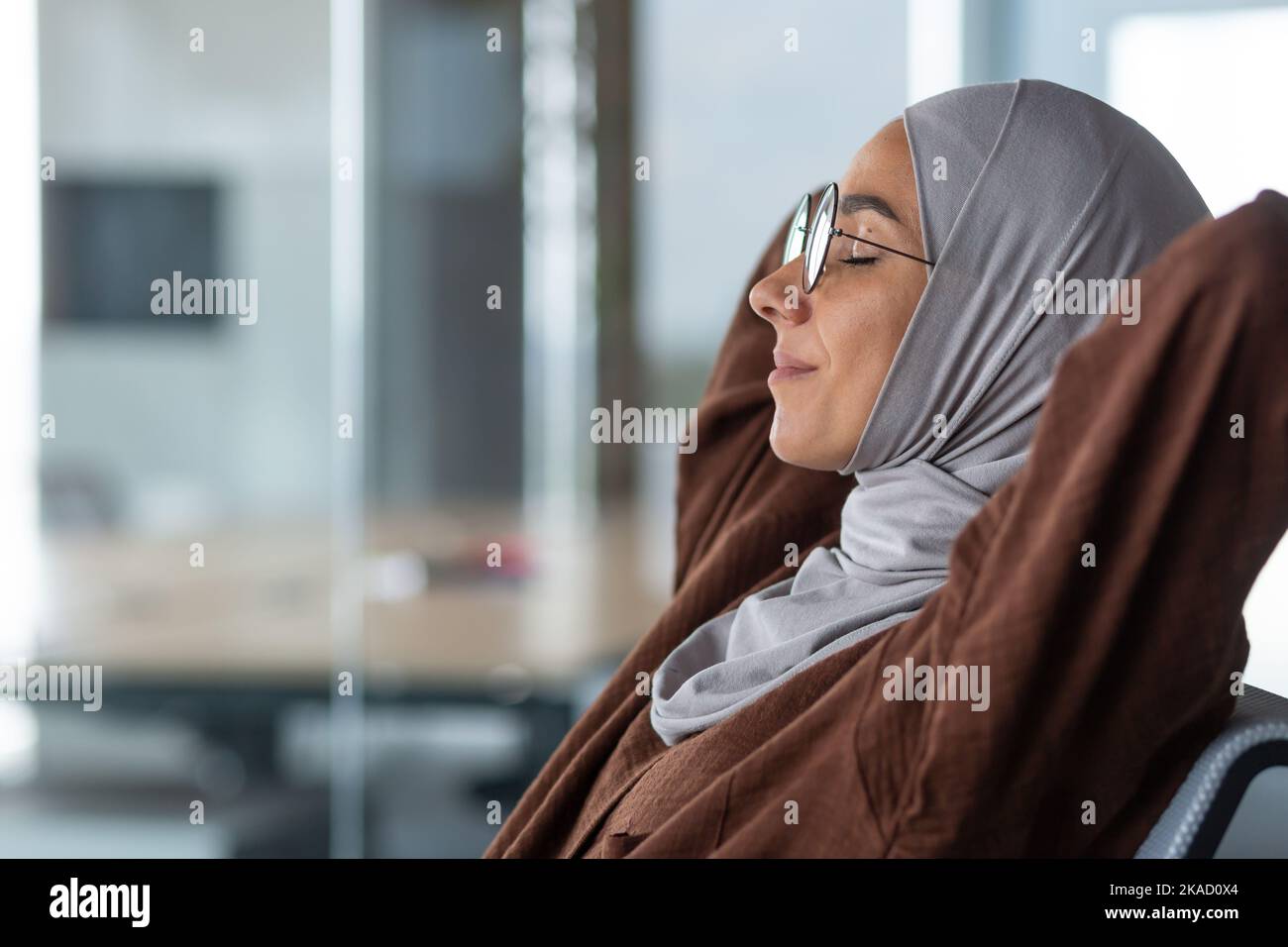 Closeup donna sognante in hijab e occhiali occhi chiusi con le mani dietro la sua testa seduta su una sedia in ufficio, donna d'affari con successo completato il suo lavoro riposante di giorno e sognante. Foto Stock