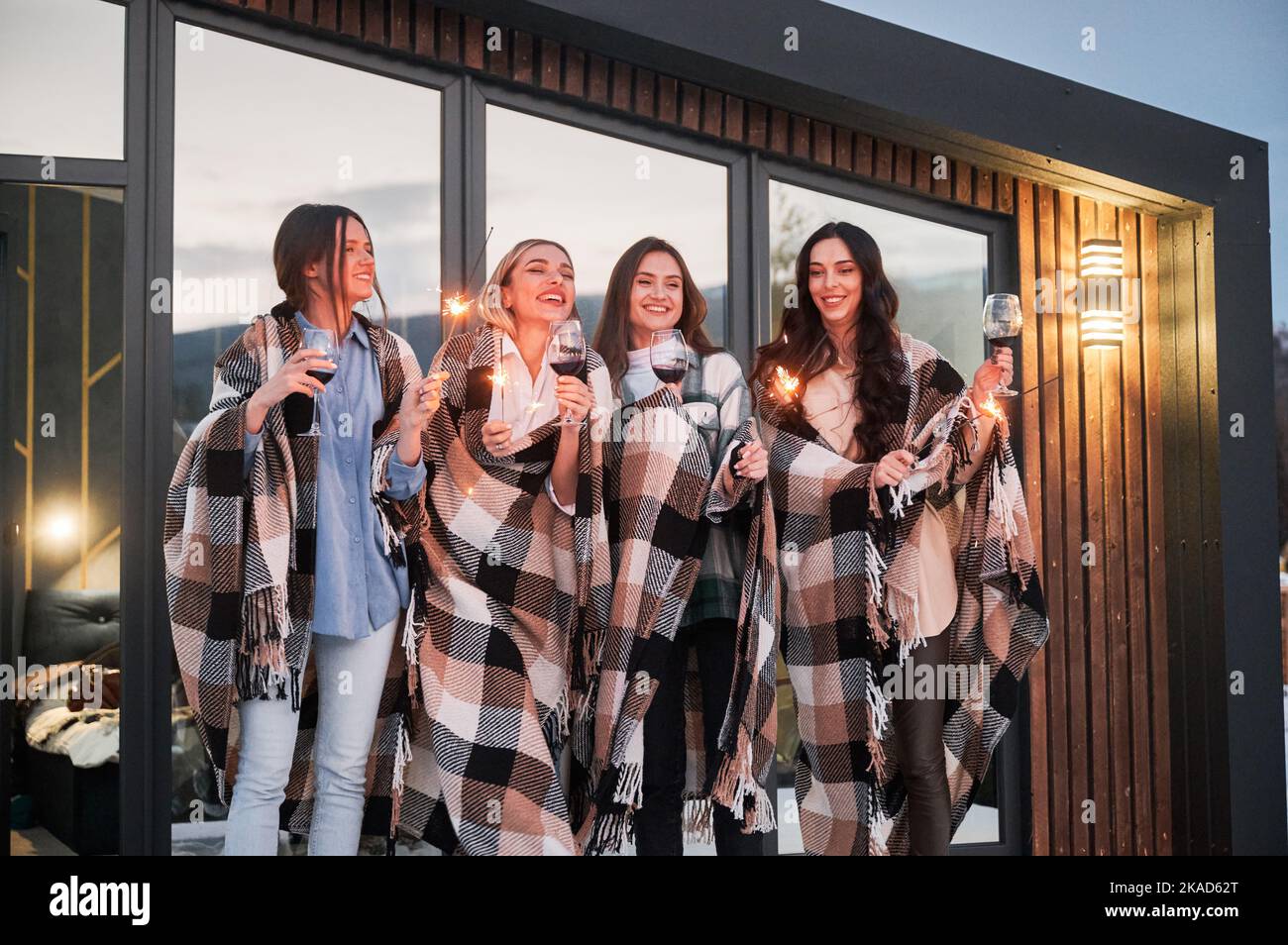 Le giovani donne che si divertiscono nei fine settimana invernali sulla terrazza della caserma contemporanea. Quattro ragazze in plaid bere vino e festeggiare con scintille la sera. Foto Stock