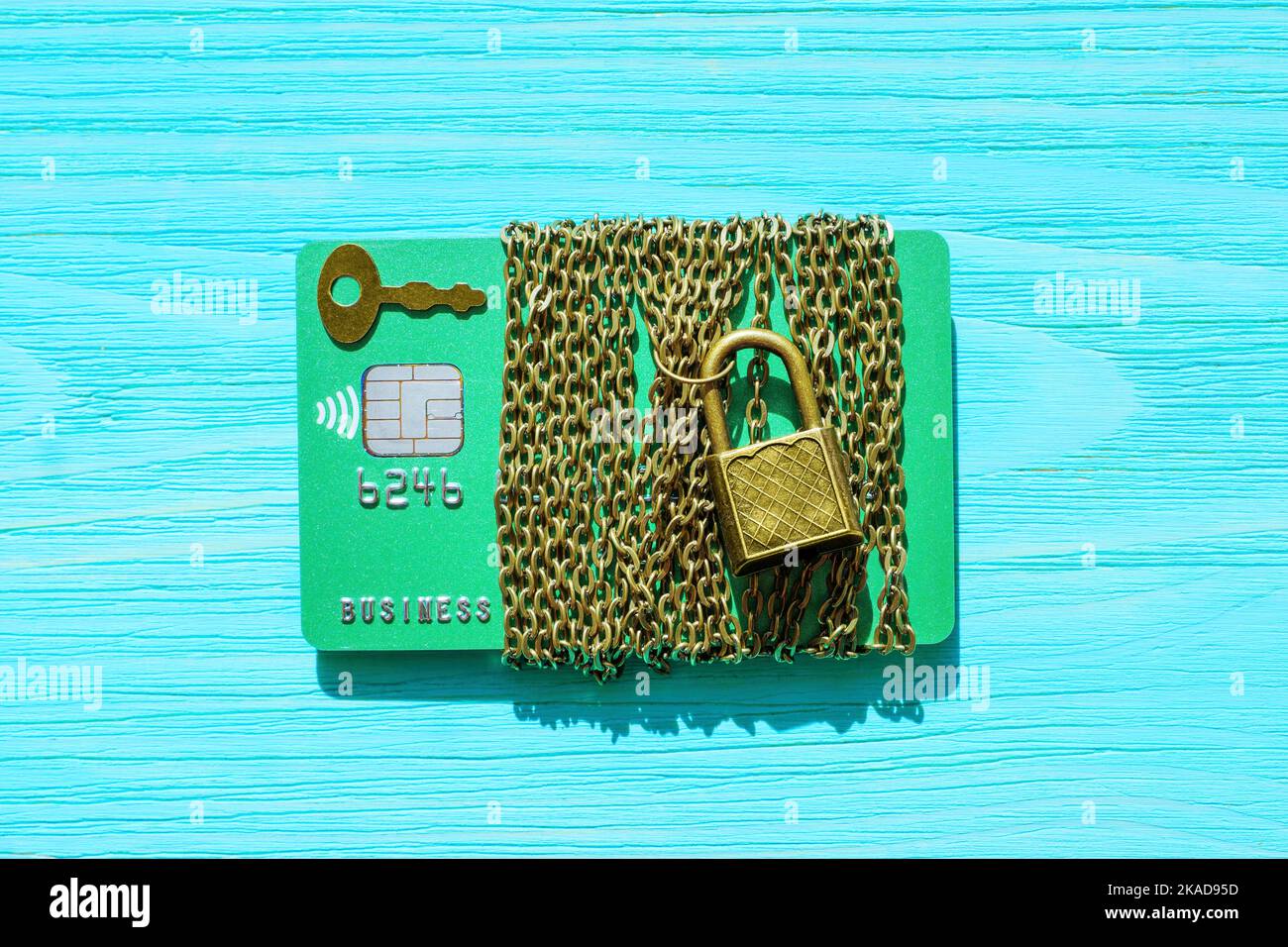 Carta di credito verde avvolta con una catena e fissata con un piccolo lucchetto isolato su uno sfondo di legno blu. Sicurezza finanziaria e protezione contro le frodi co Foto Stock
