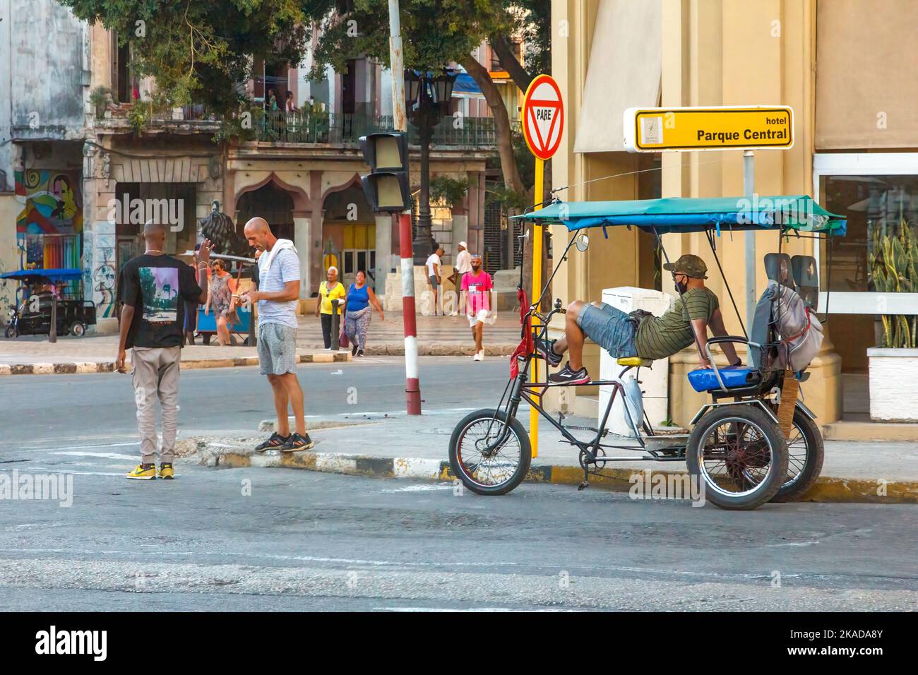 Un conducente bicitaxi o pedicab si appoggia all'indietro mentre attende i clienti. Altri cubani si trovano all'angolo della città. Foto Stock