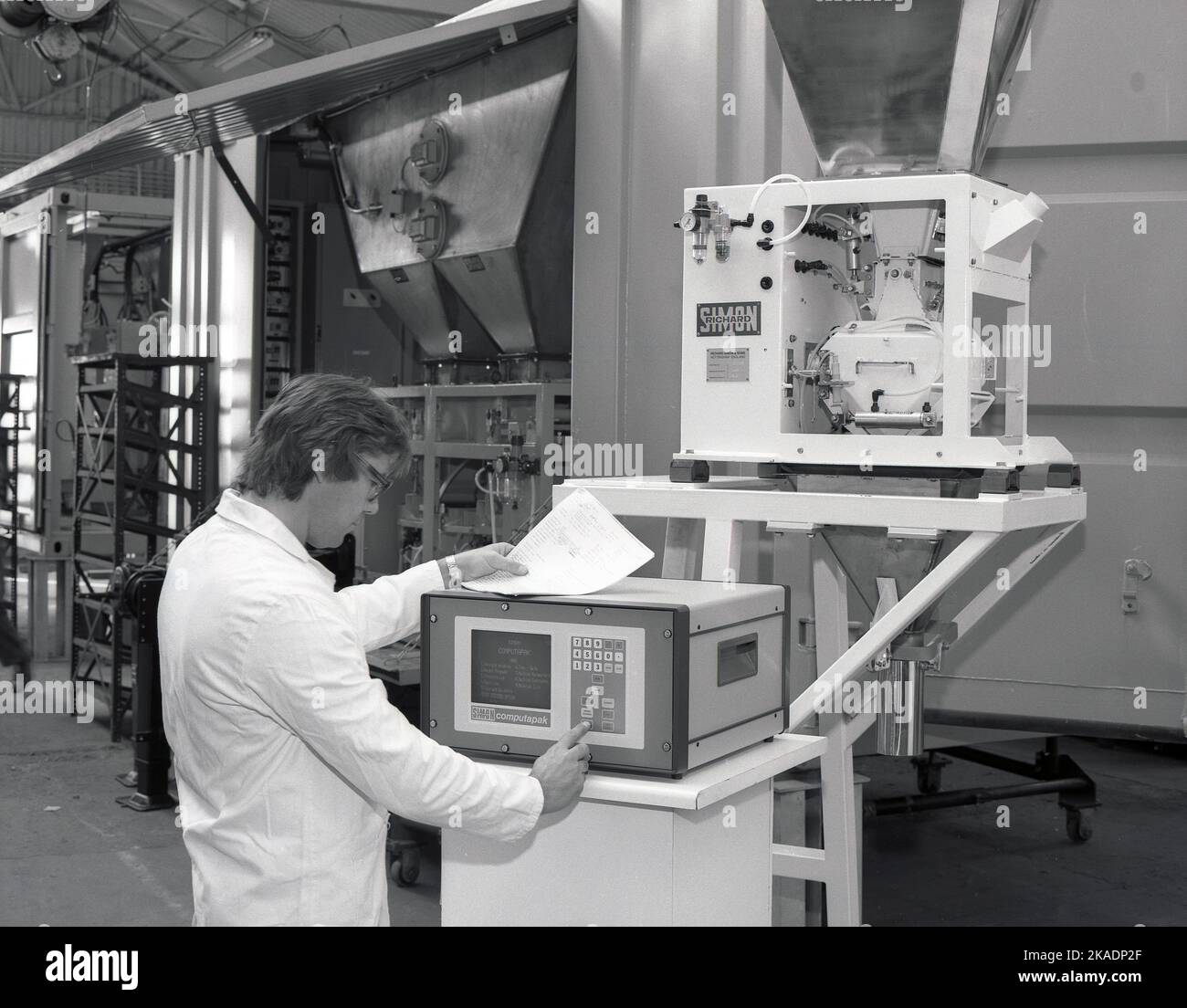 1980s, storico, tecnico maschile che aziona il computer di una macchina su una macchina riempitrice di fertilser, fatta da Richard Simon & Sons, di Nottingham, Inghilterra, UK. Schermo sulla macchina dice Simon Computapak, con menu che dà opzioni compreso il peso Foto Stock