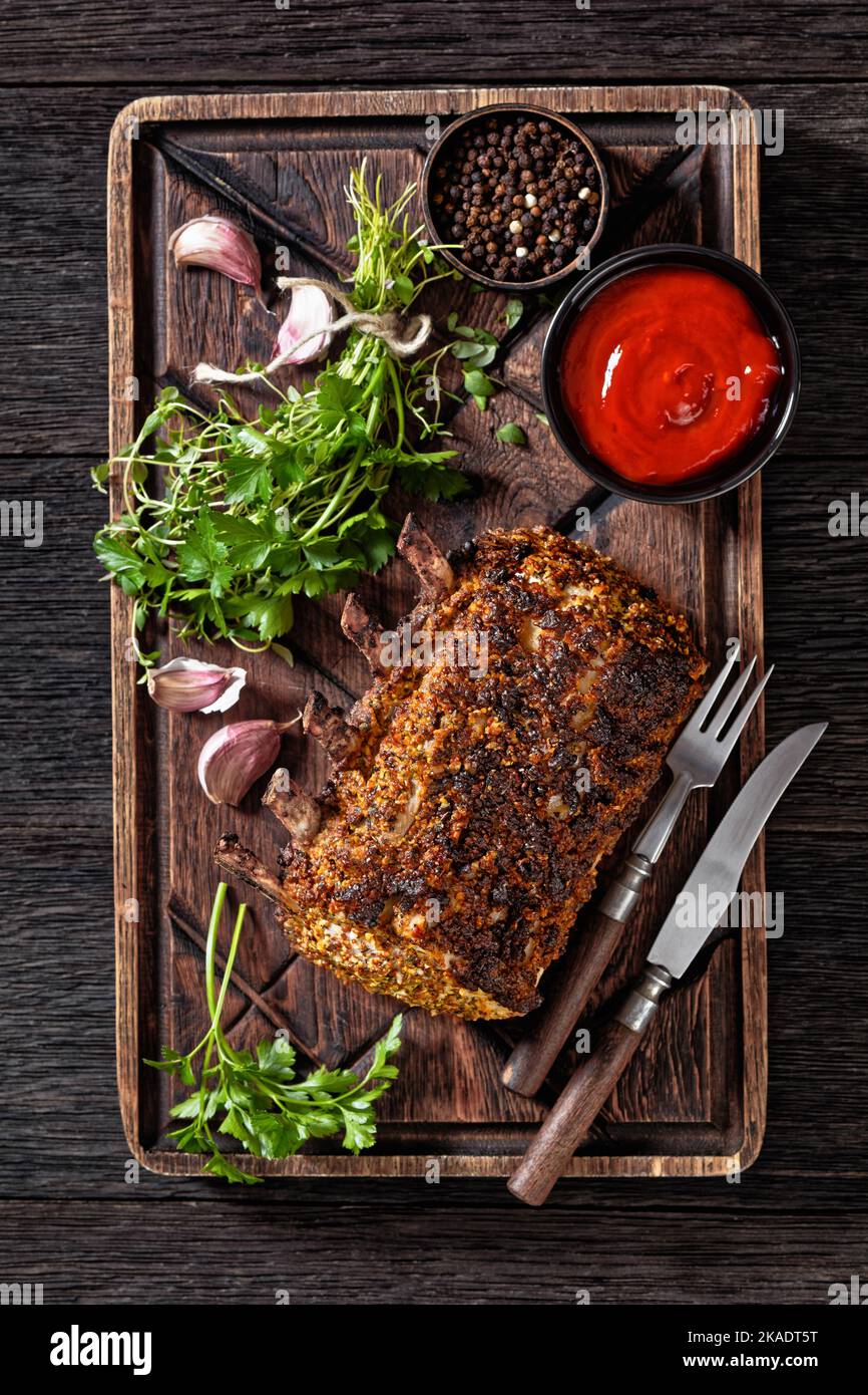 Costolette di maiale in piedi arrosto su un tagliere rustico con posate, salsa di pomodoro, timo e prezzemolo, vista verticale dall'alto Foto Stock