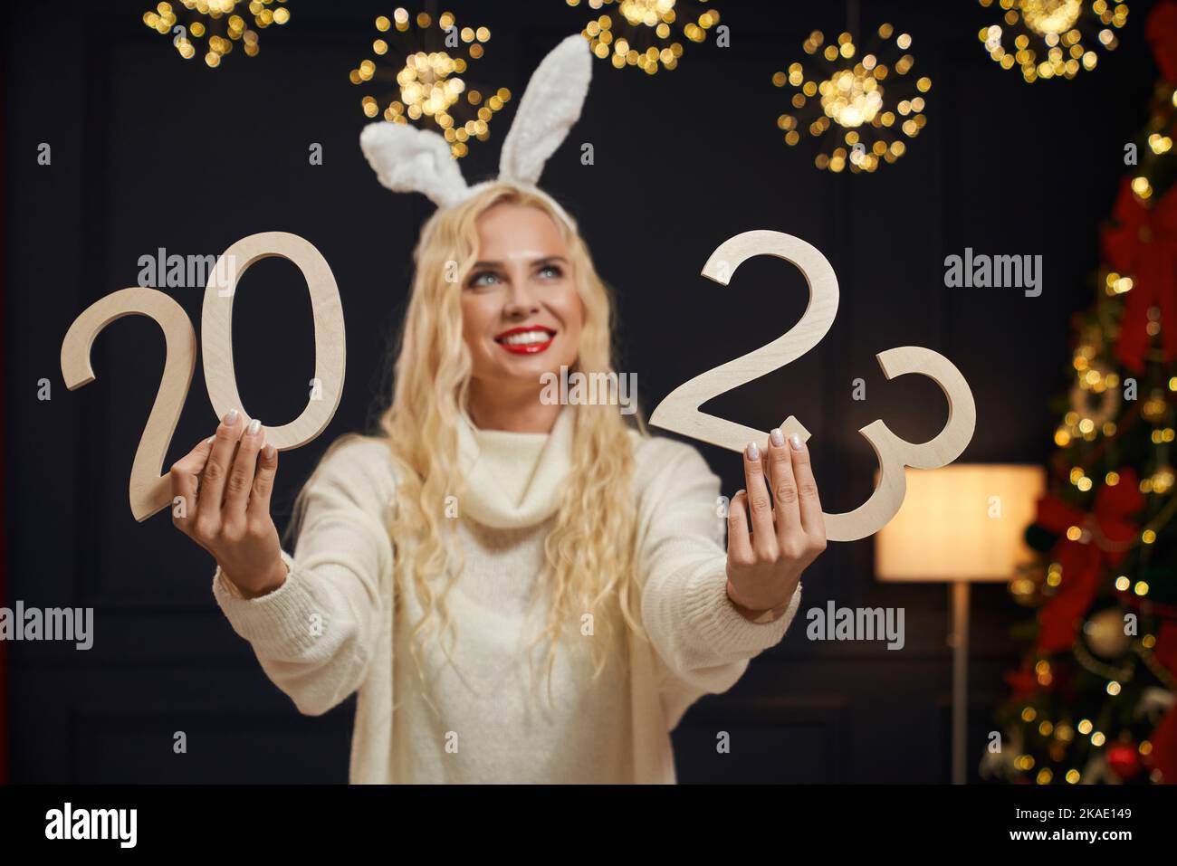 Vista frontale di una bella donna che indossa un maglione a maglia, guardando in alto, tenendo 2023 decorazione, sorridendo. Bella ragazza bionda con labbra rosse in piedi, sognando. Concetto di nuovo anno e Natale. Foto Stock