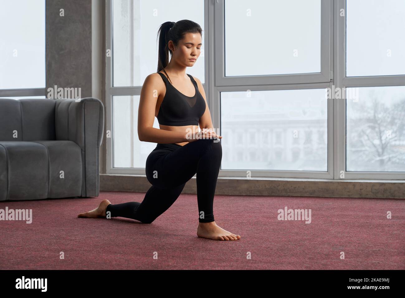 Vista laterale della ragazza brunetta in piedi su un ginocchio con gli occhi chiusi all'interno. Giovane donna sportiva che pratica yoga, meditando, facendo un passo avanti. Concetto di nuova era realtà. Foto Stock