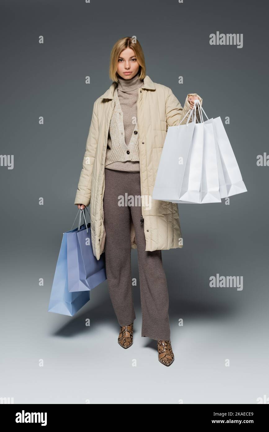 lunghezza intera di giovane donna bionda in cappotto puffer di inverno che tiene i sacchetti di shopping sul grigio, immagine di riserva Foto Stock