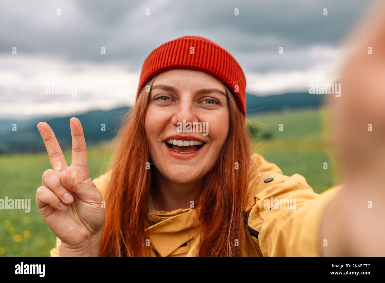 Giovane escursionista sorridere felice donna in una giacca gialla e un cappello rosso brillante che prende selfie ritratto sulla cima della montagna, mostra gesto di pace. Escursioni Foto Stock