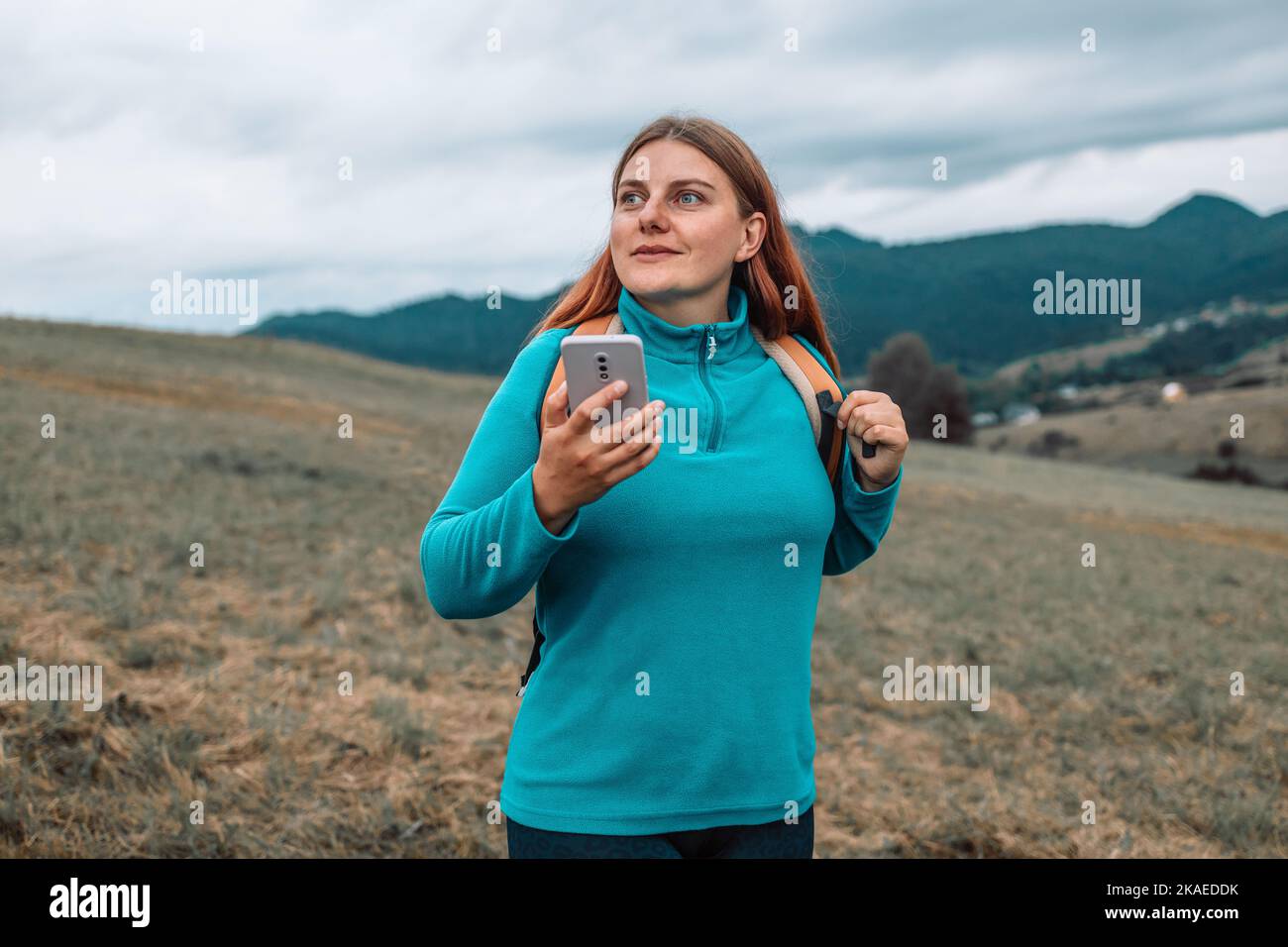 Una donna con uno zaino si erge sulla cima di una montagna utilizzando smartphone e ammira la bellezza di una valle di montagna, una vista posteriore. Foto Stock