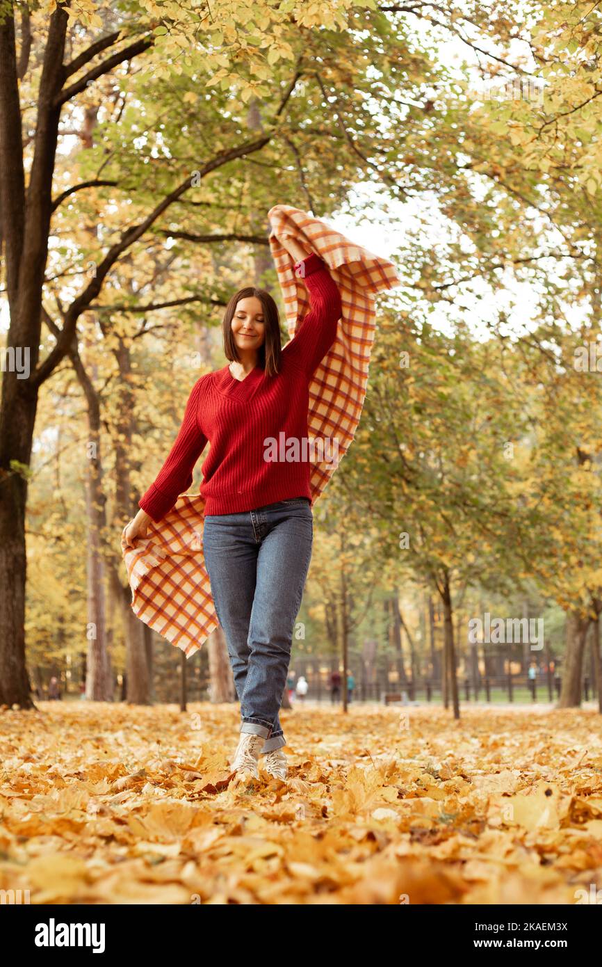 Foto a tutta lunghezza di felice donna allegra in confortevole maglione rosso lavorato a maglia divertirsi con la plaid a scacchi, fogliame dorato sul terreno. Giovane donna entrare Foto Stock