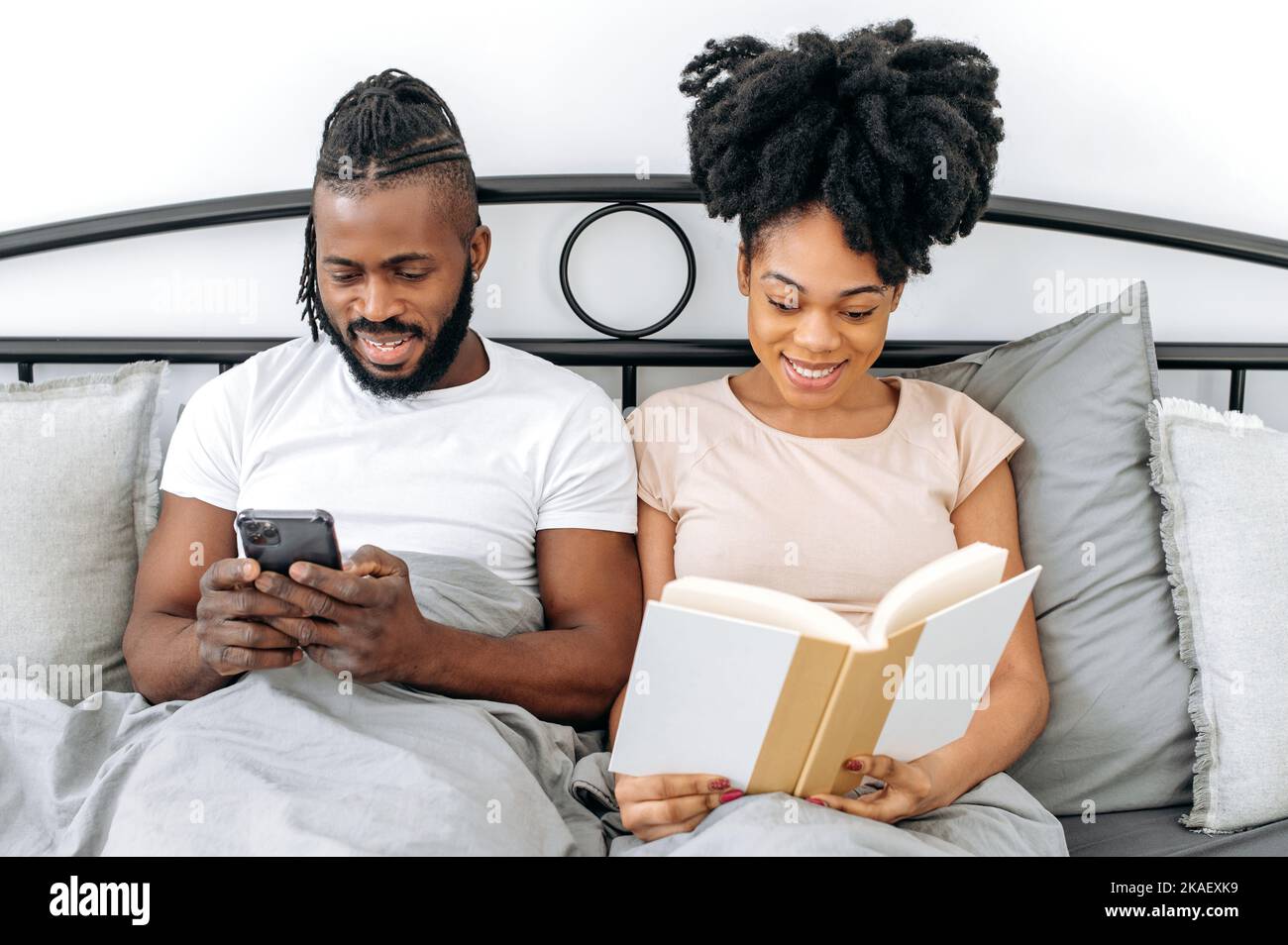 Sposi felici, uomo e donna afro-americana, sono seduti a letto nella camera da letto, l'uomo sta usando uno smartphone, leggendo le notizie, navigando in Internet, la donna che legge un libro interessante prima di andare a letto Foto Stock