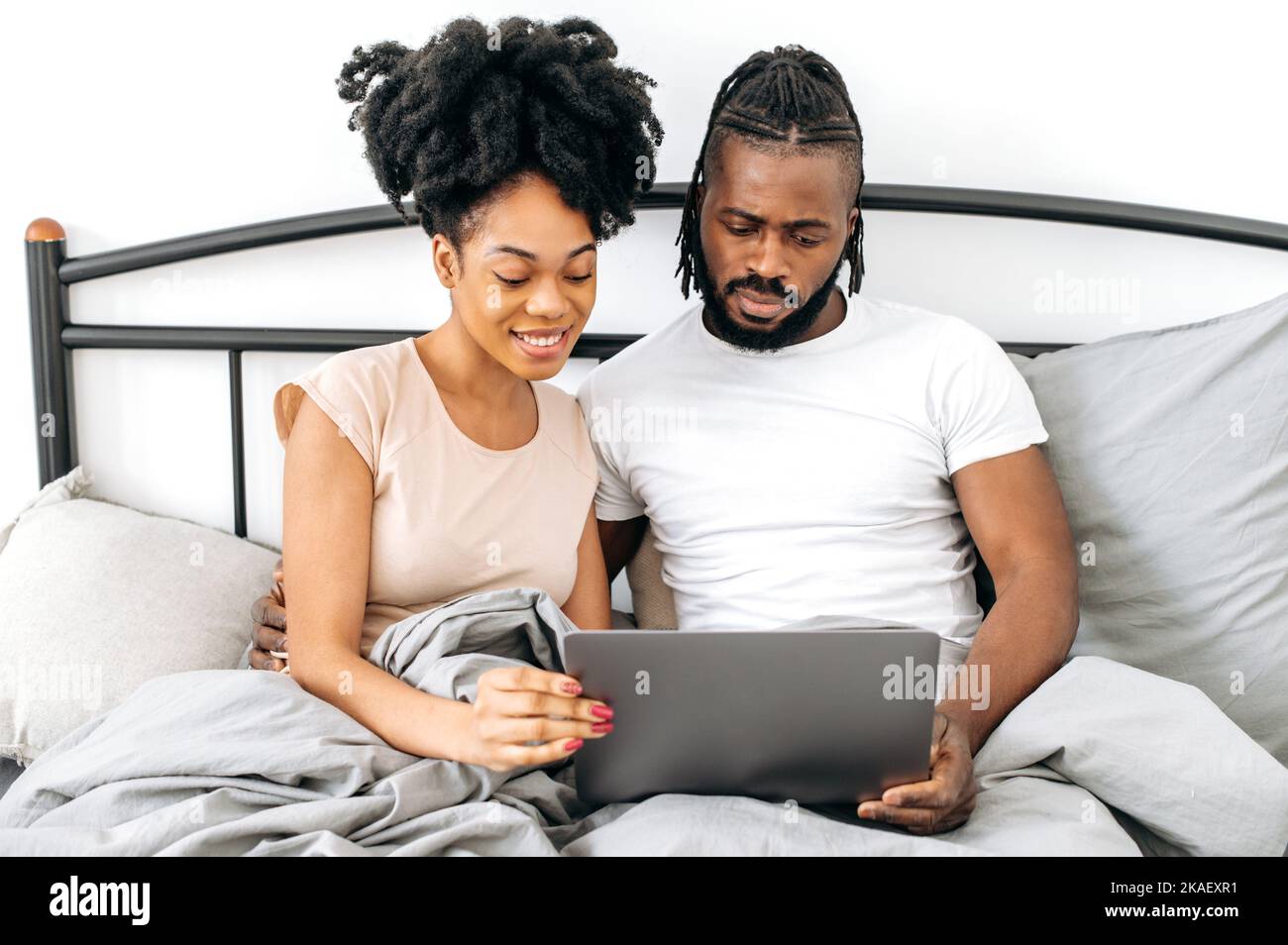 Sposi afro-americani, marito e moglie, positivi e soddisfatti, sono seduti in un letto in camera da letto, utilizzando il laptop, navigando in internet, guardando film insieme prima di andare a letto, leggendo notizie, sorridendo Foto Stock