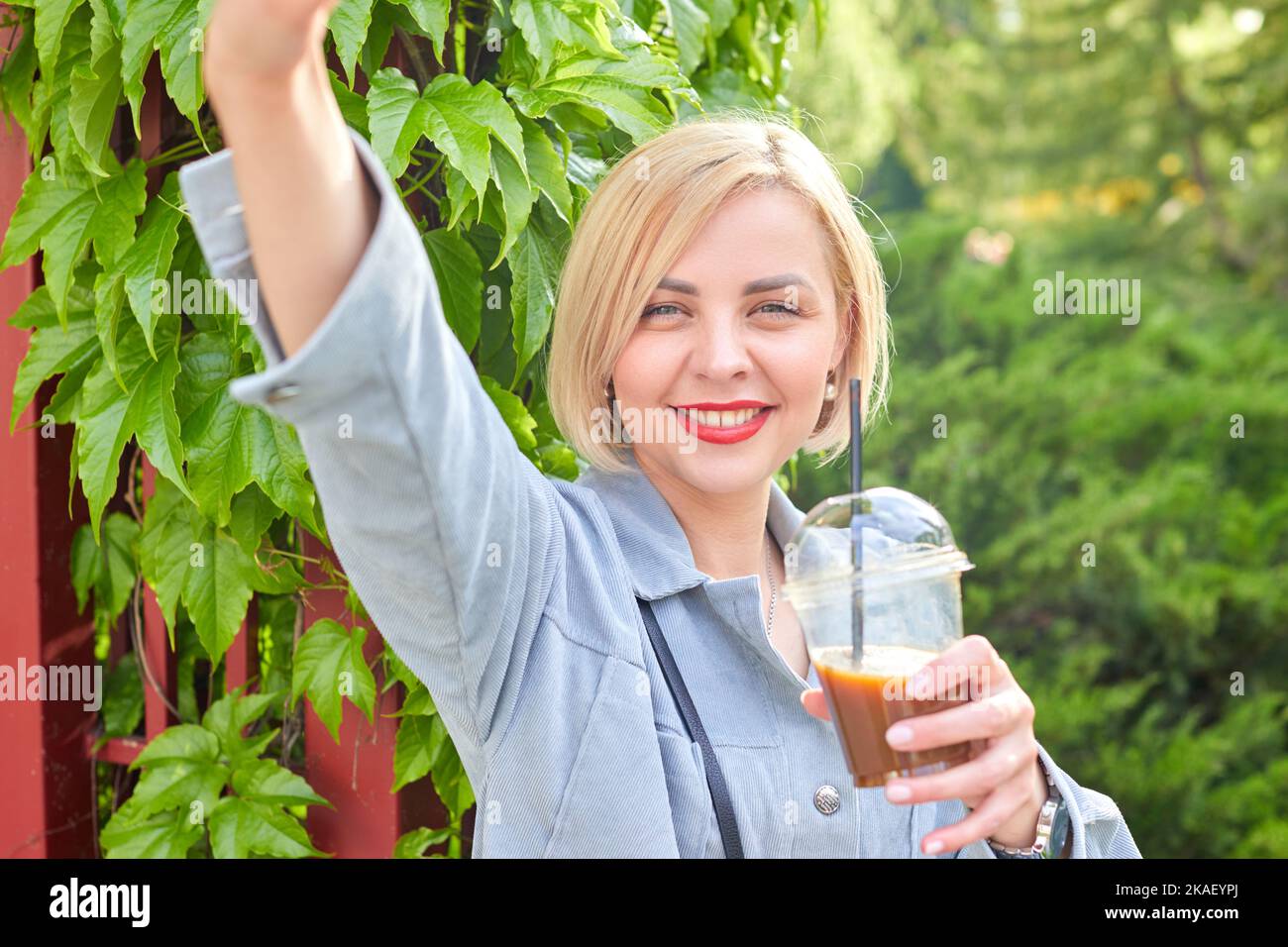 Elegante giovane bionda cammina per la città, beve una bevanda fredda e prende un selfie sul suo telefono cellulare. Tempo trascorso dopo il lavoro Foto Stock