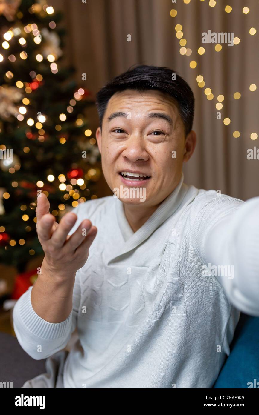 Giovane asiatico che parla sulla fotocamera del telefono come selfie. Si siede a casa vicino all'albero di Natale, saluta, comunica, sorride. Foto Stock