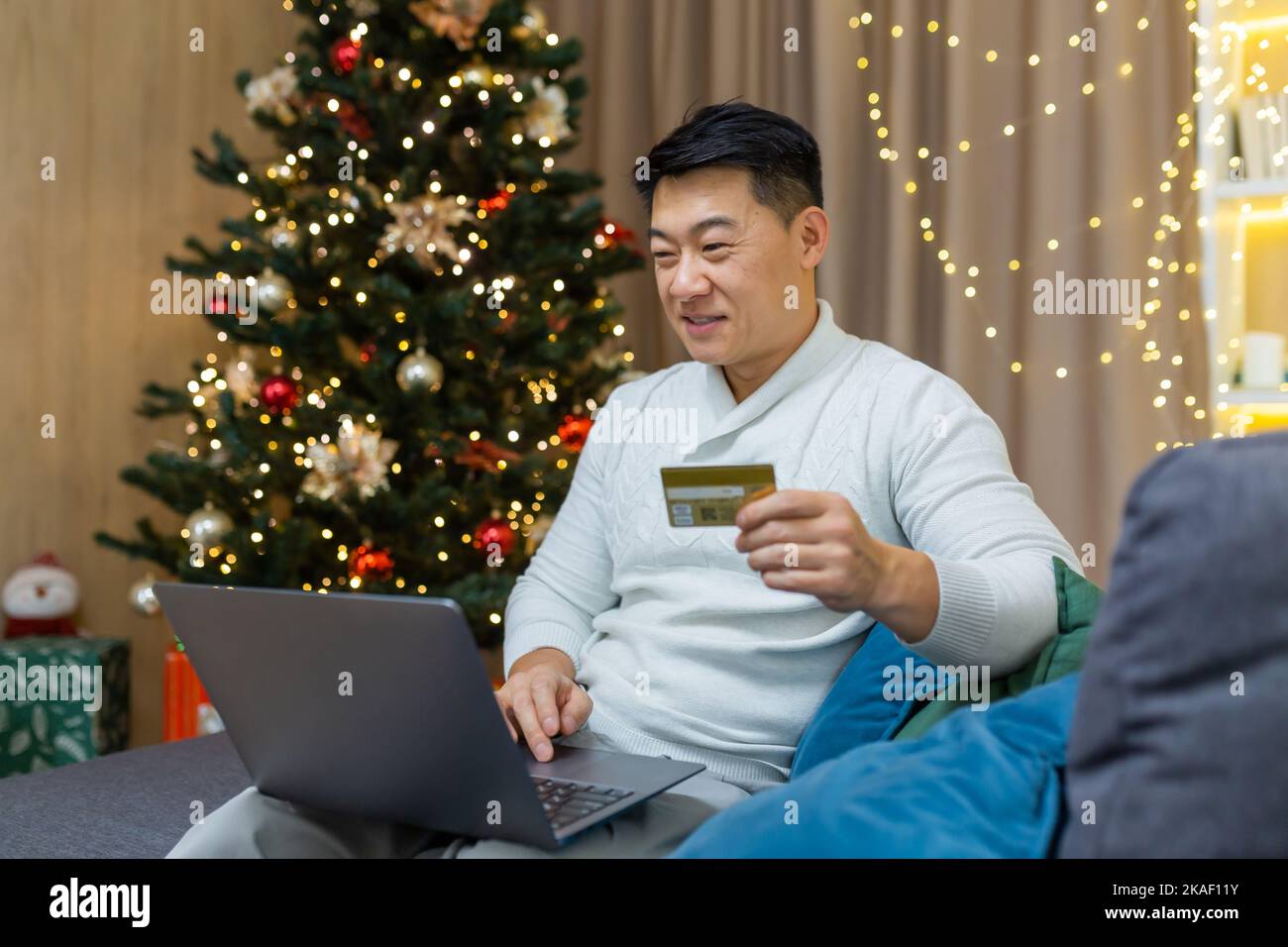 Shopping online festivo. Un giovane asiatico è seduto sul divano vicino all'albero di Natale a casa. Ha un computer portatile e una carta di credito. Shopping online per regali di Natale per la famiglia e gli amici. Foto Stock