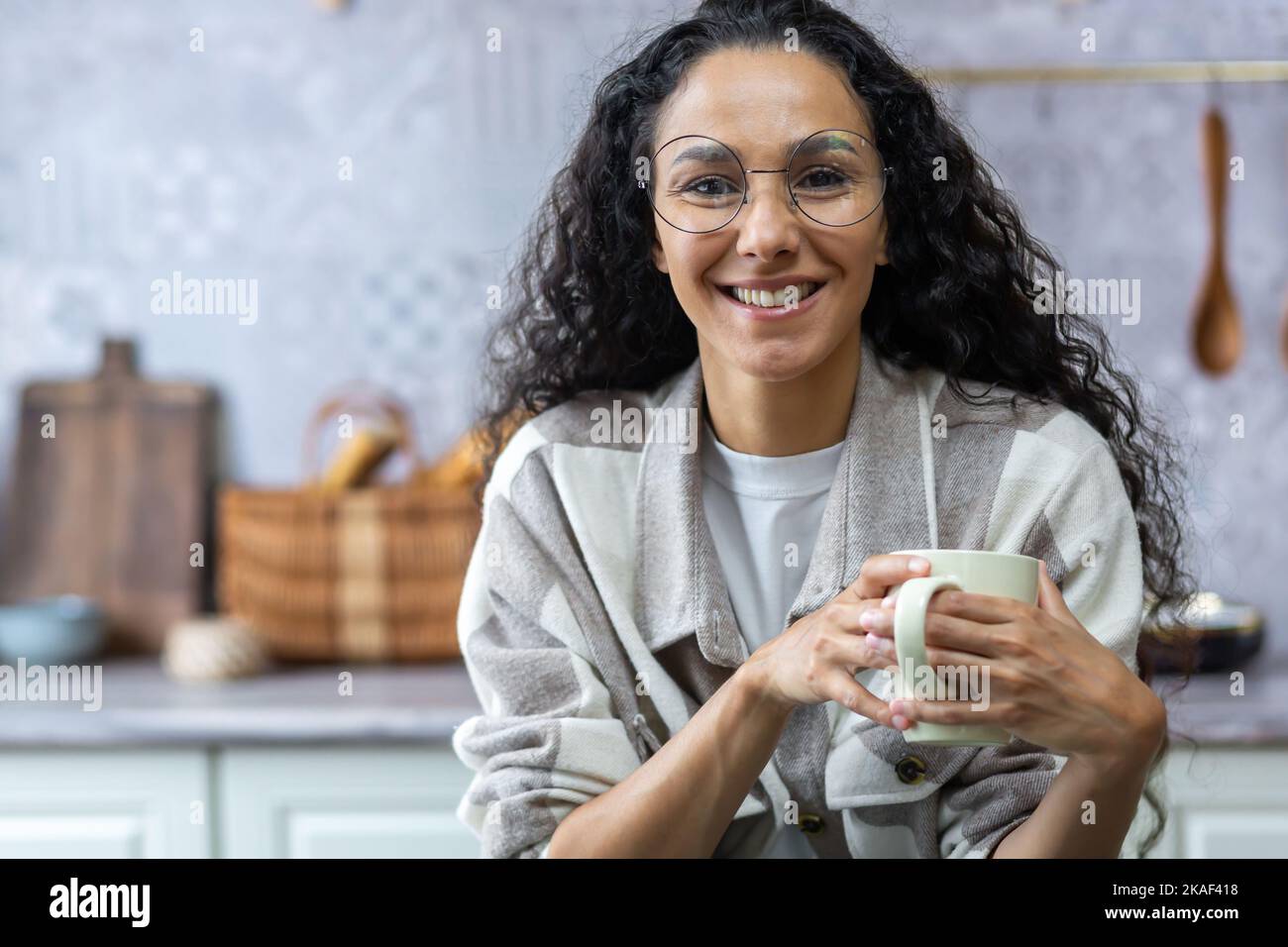 Videochiamata, comunicazione online con gli amici, felice e sorridente donna ispanica guardando la webcam e parlando, donna a casa in cucina con capelli ricci e bicchieri che tiene una tazza di bevanda calda. Foto Stock