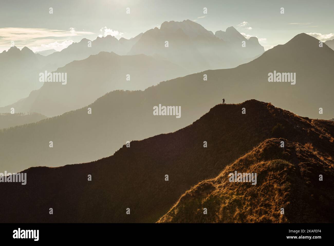 Un uomo si erge su una cima panoramica del Passo di Giau nella calda retroilluminazione di fronte alle catene montuose e al Monte Marmolada, Dolomiti, Italia Foto Stock