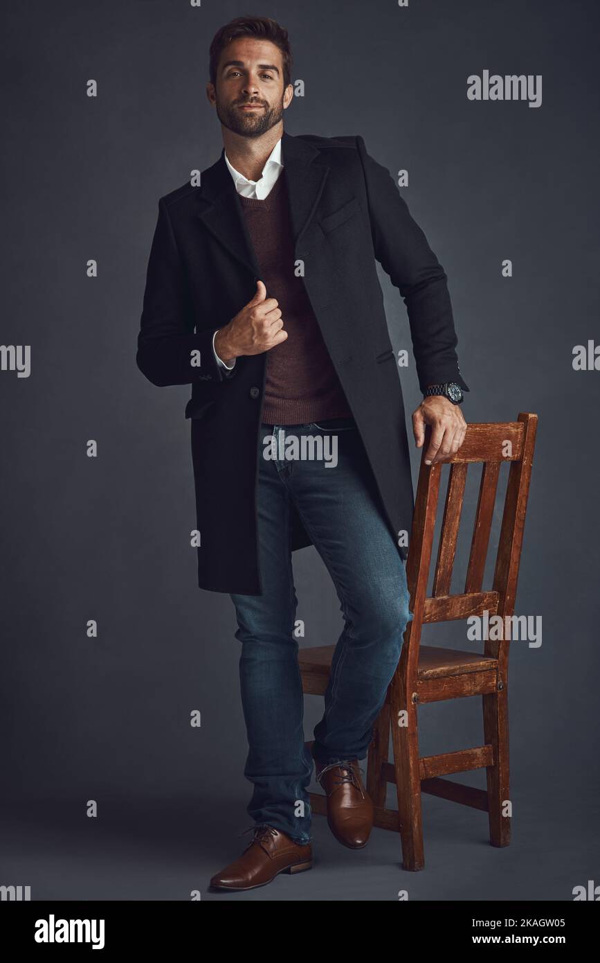 Obiettivi moda. Studio ritratto di un giovane uomo elegantemente vestito in piedi accanto a una sedia su uno sfondo grigio. Foto Stock
